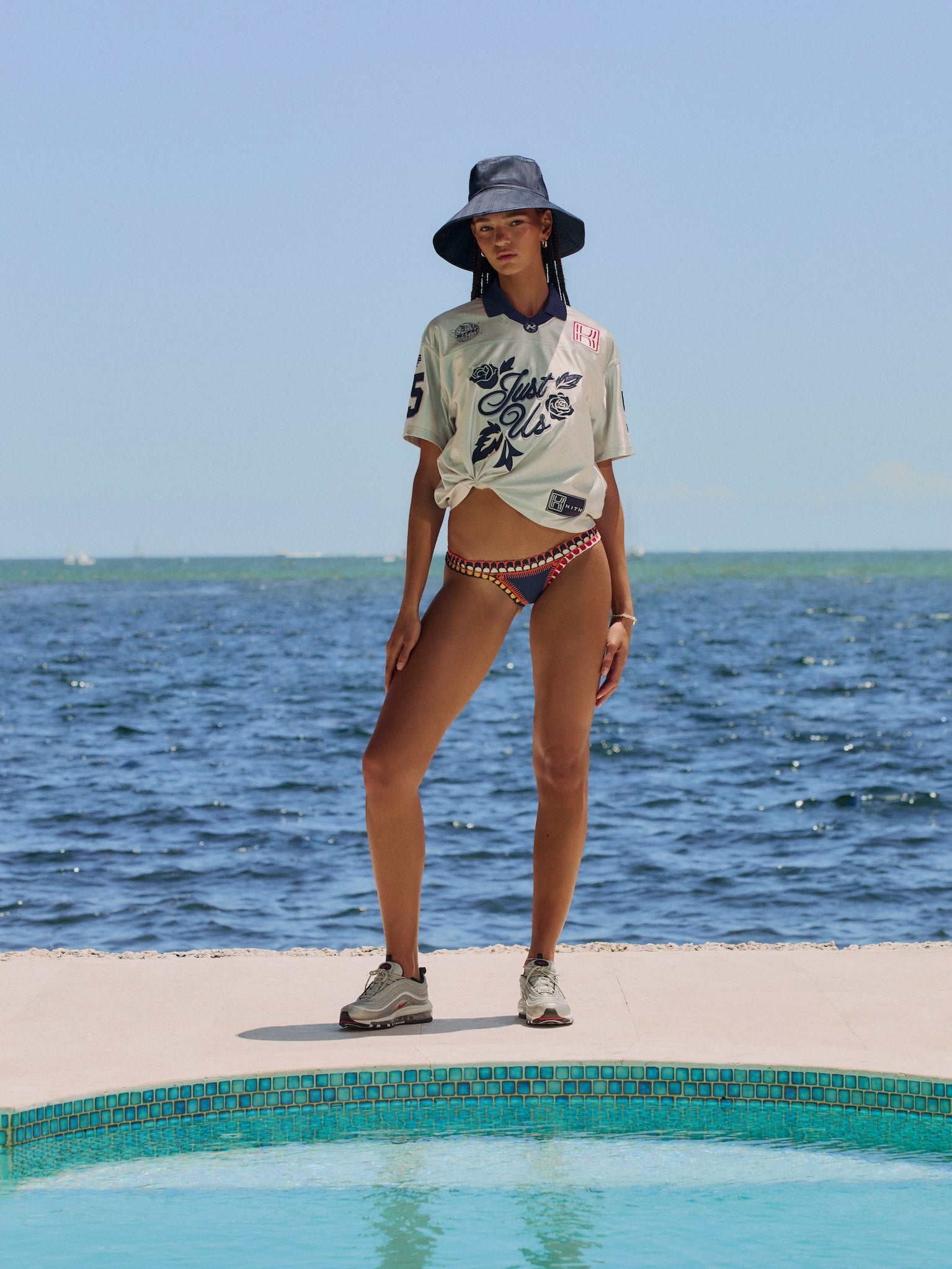 Model at an oceanfront pool in a navy bucket hat and knotted cream rose-print jersey with bikini bottoms, Nike Air Max 97