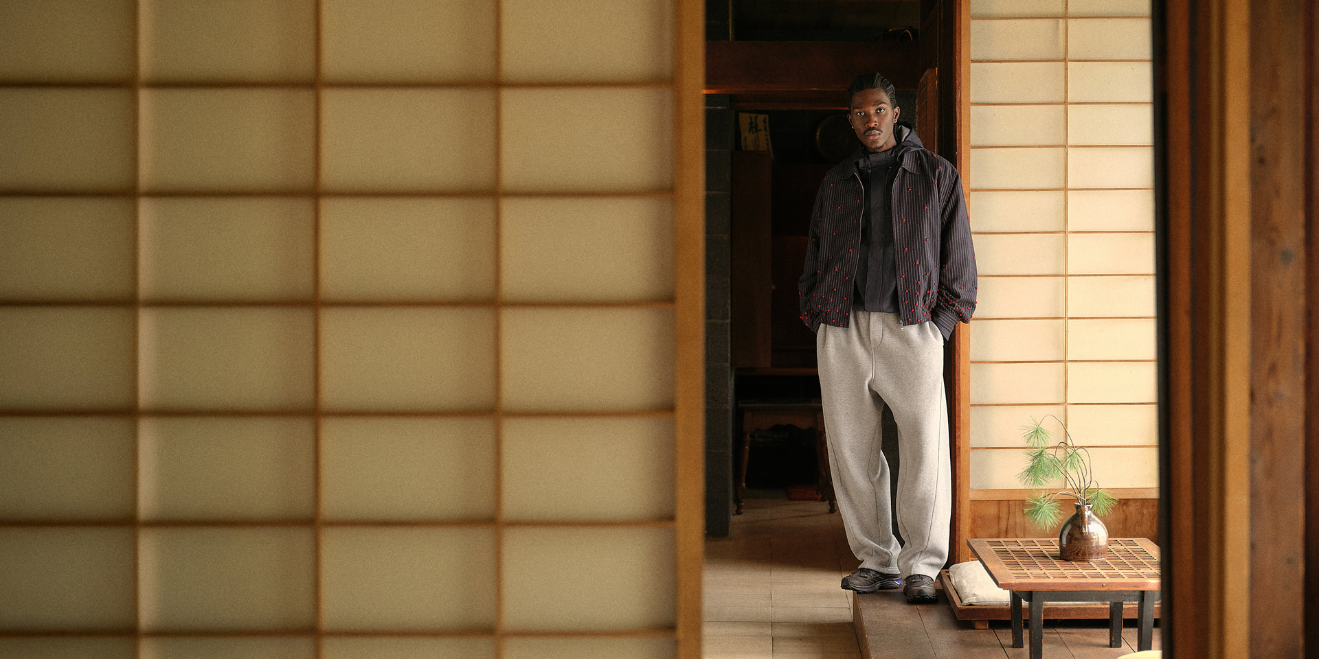 Man in navy pinstripe jacket, black hoodie and grey wide-leg sweatpants in a Japanese shoji-screen room