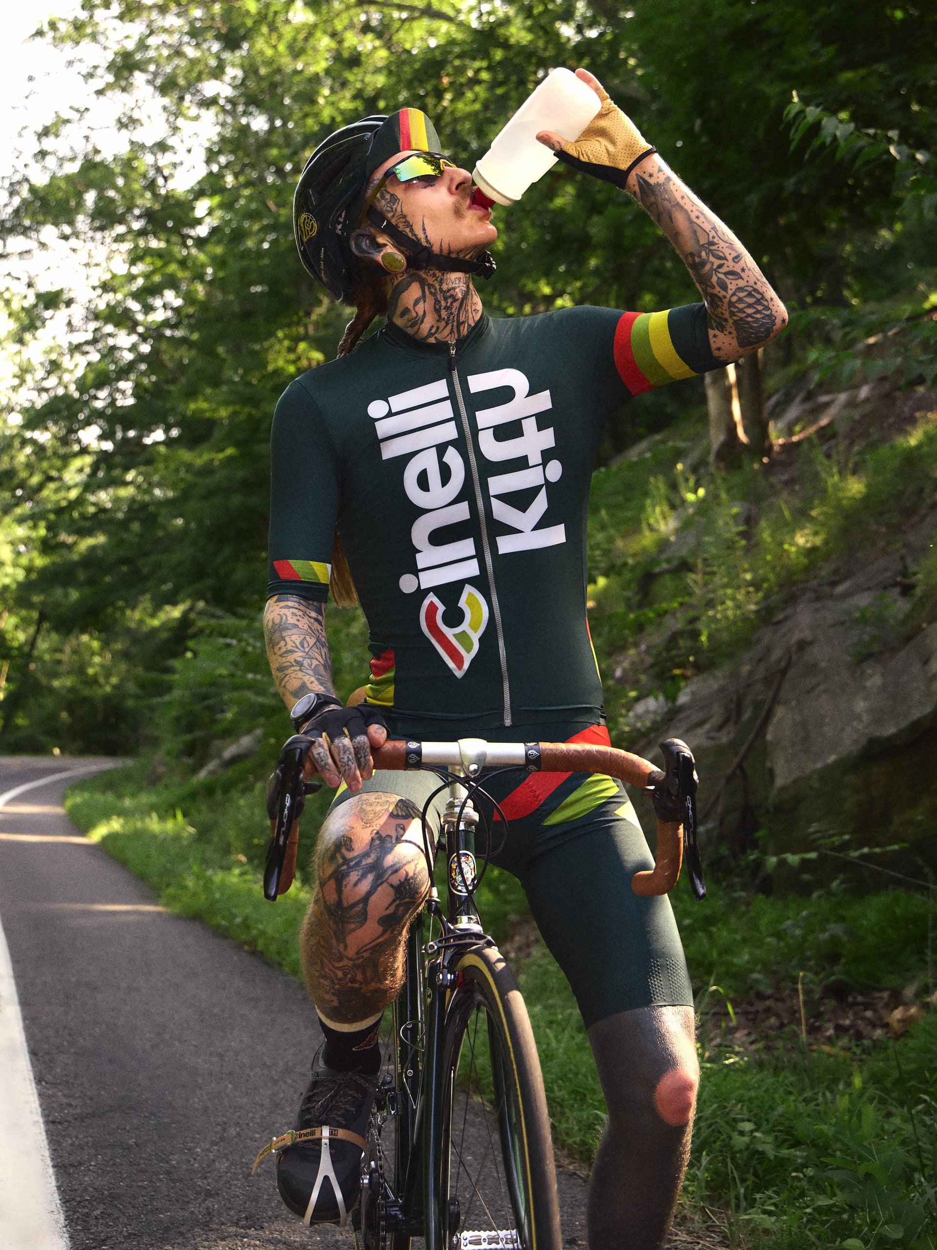 Tattooed cyclist drinking from a bottle in a green Cinelli x Kith cycling jersey on a forest road