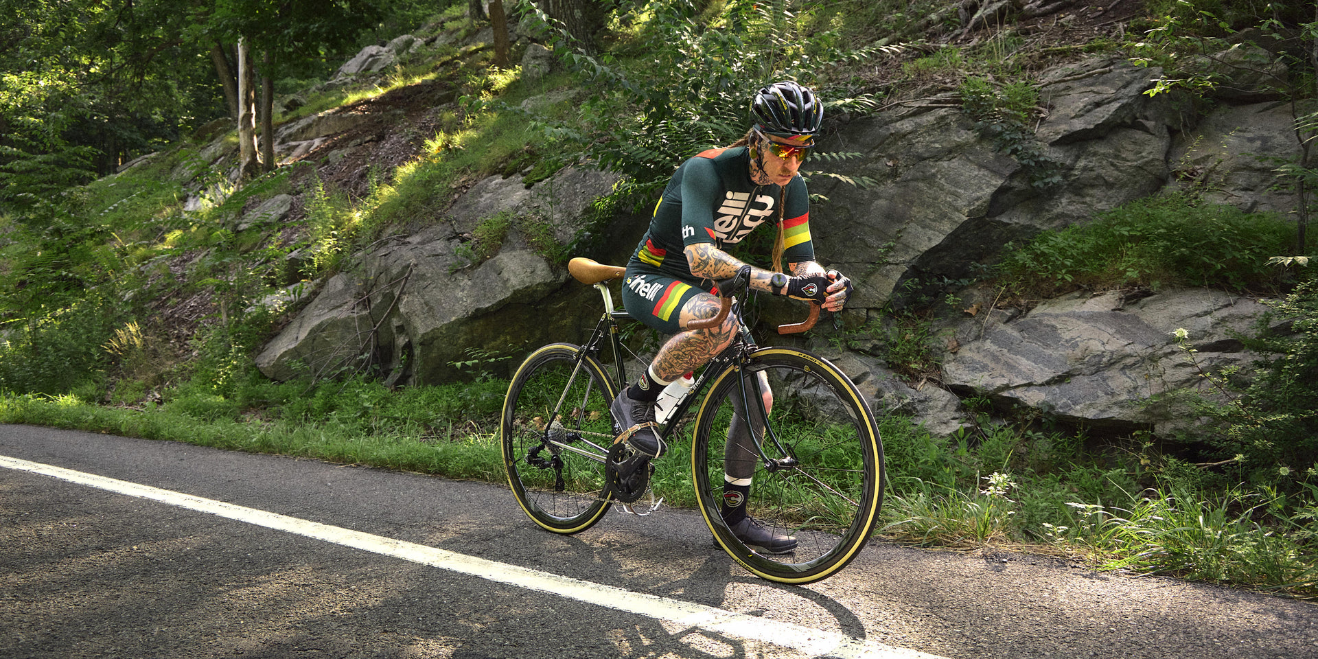 Cyclist riding a road bike in green Cinelli x Kith kit along a rocky forest road, wide view
