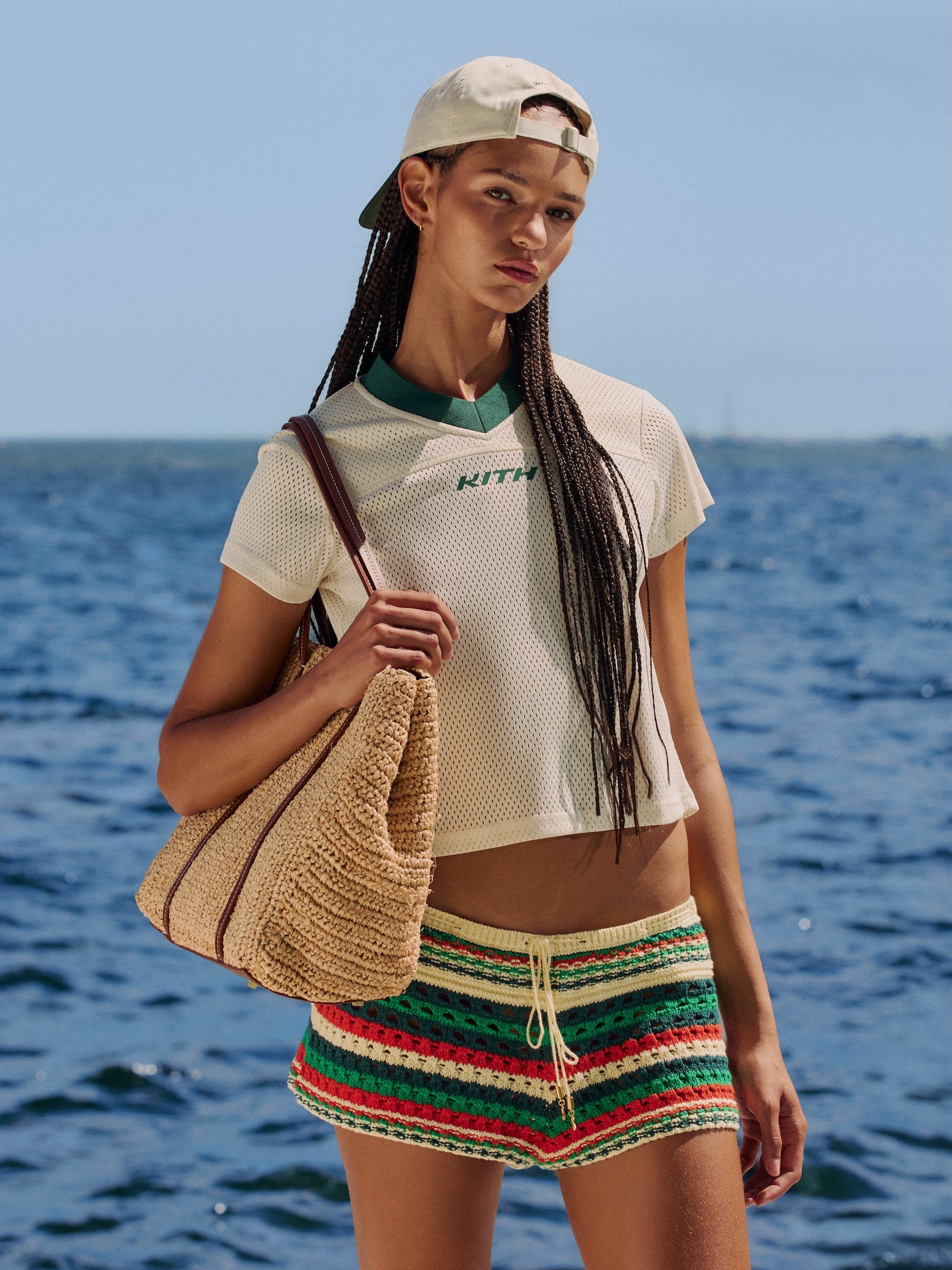 Model by the sea in a cream KITH mesh V-neck tee, crochet striped shorts and a backwards cap, carrying a woven raffia tote