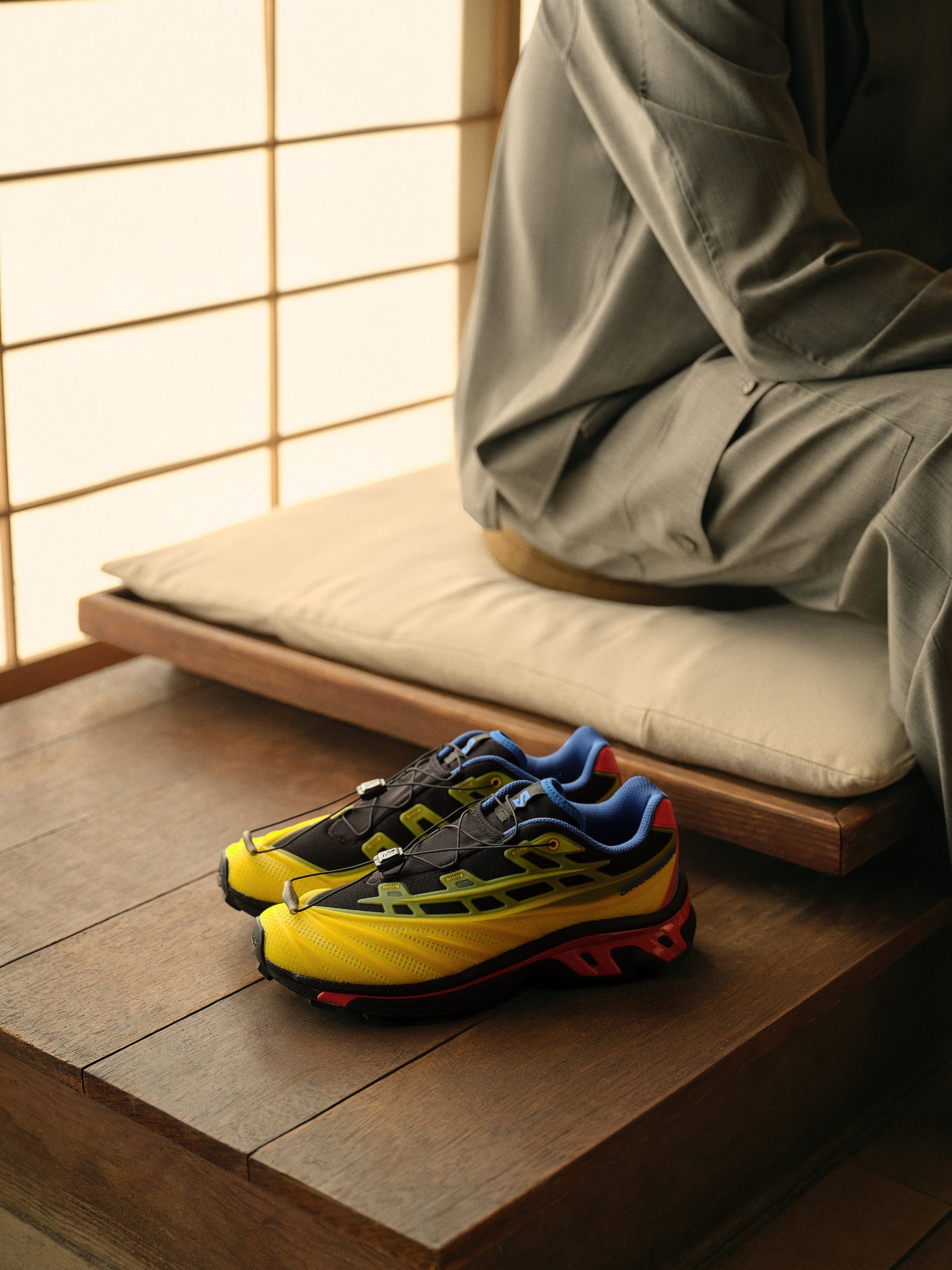 Yellow, blue and red Salomon XT sneakers on a wood bench beside a person in grey trousers