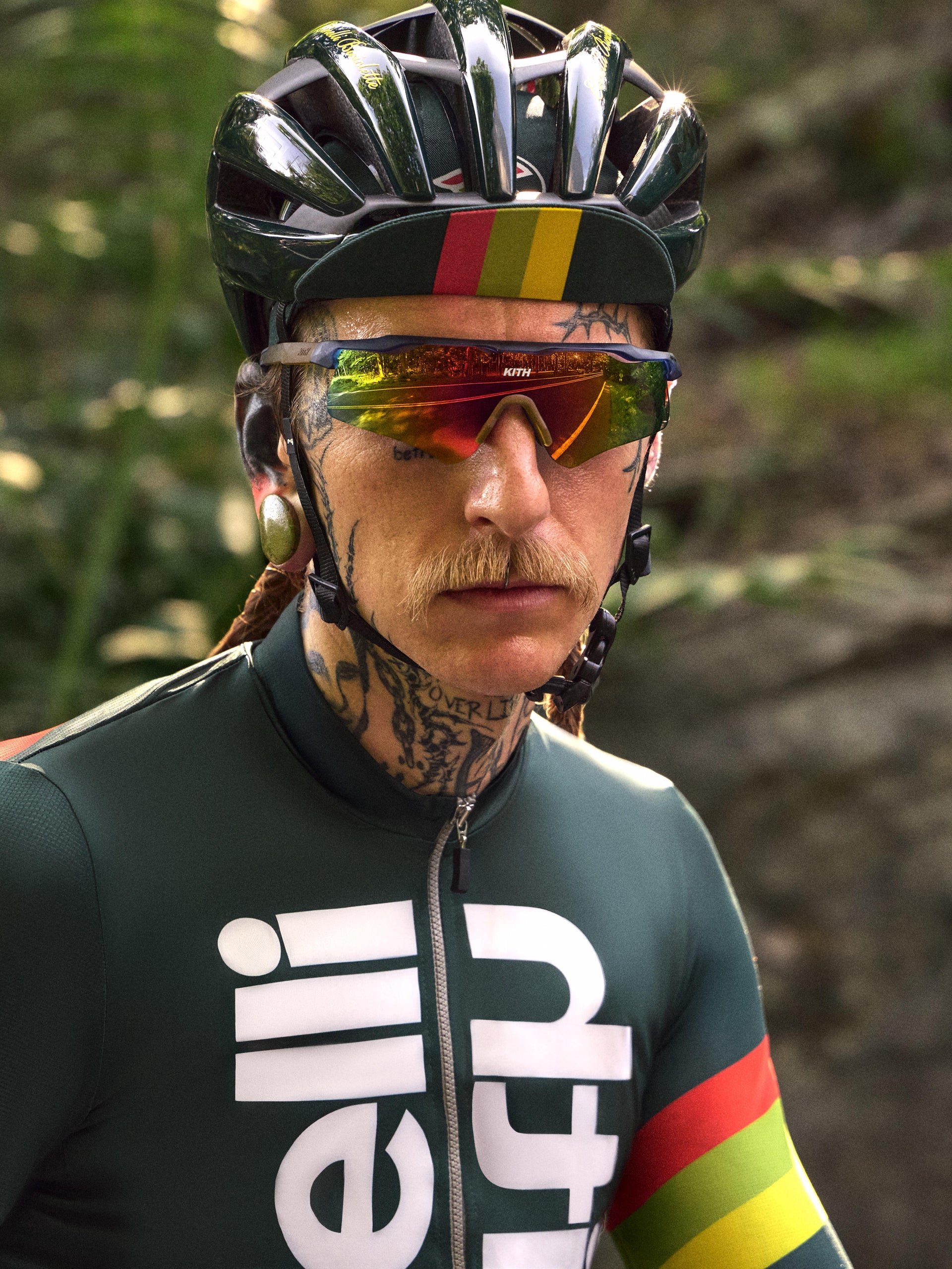 Close-up portrait of a tattooed cyclist in a green Cinelli x Kith jersey, helmet and Kith sunglasses