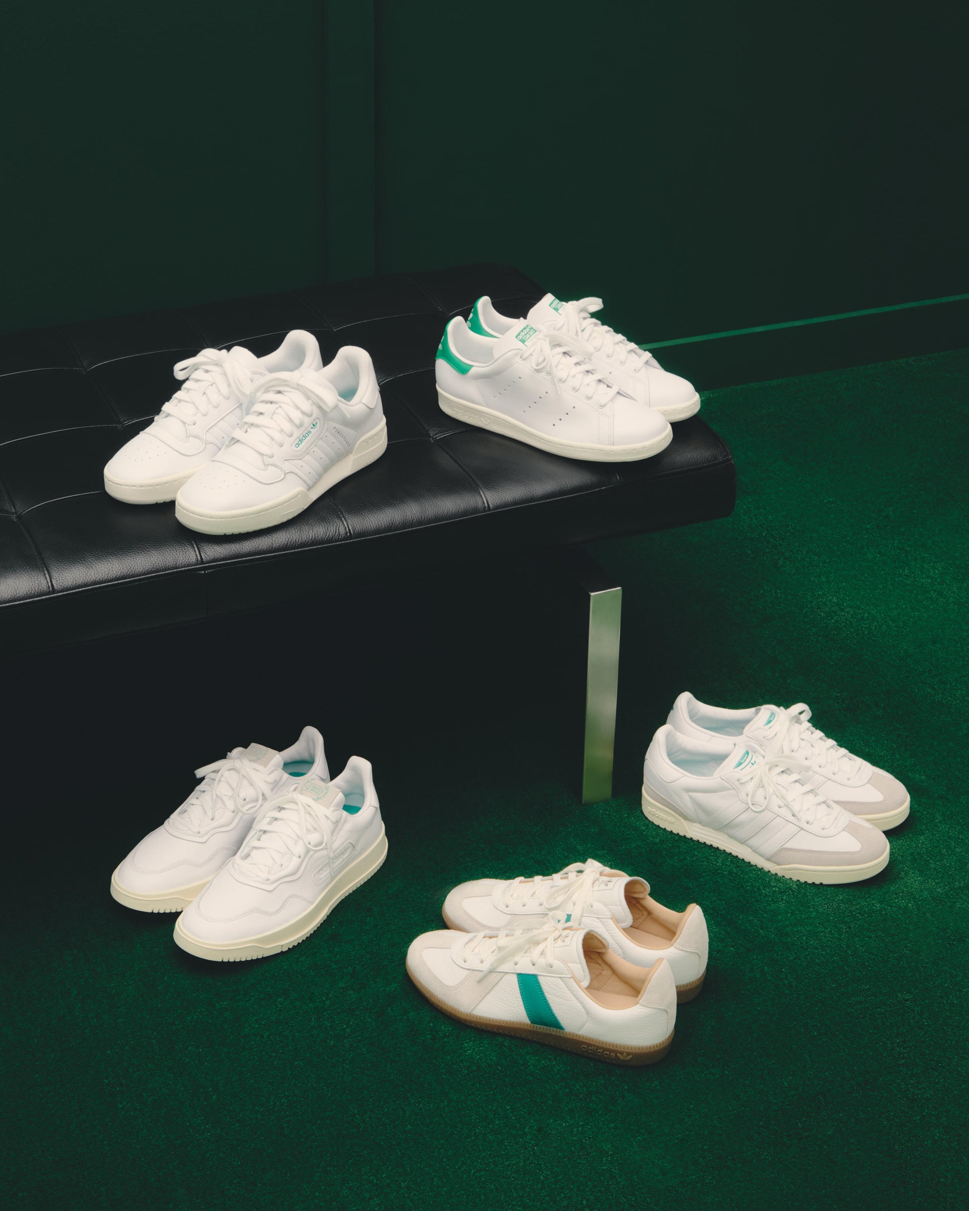 White adidas and Kith low-top sneakers with green accents arranged on a black leather bench and green studio carpet