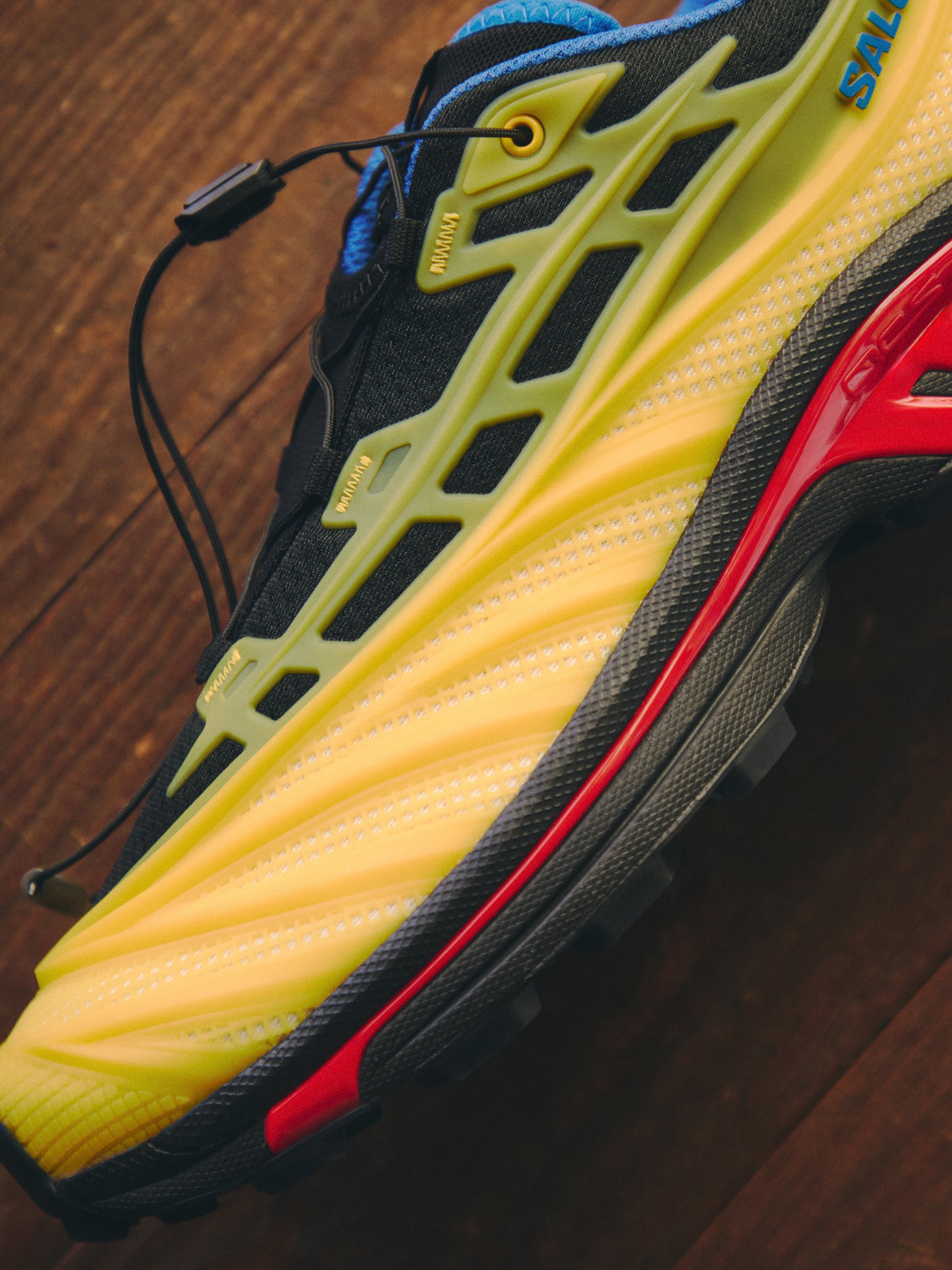 Close-up of Salomon XT-6 trail sneaker in yellow, black and red with red midsole, mesh upper and quicklace, on a wood floor