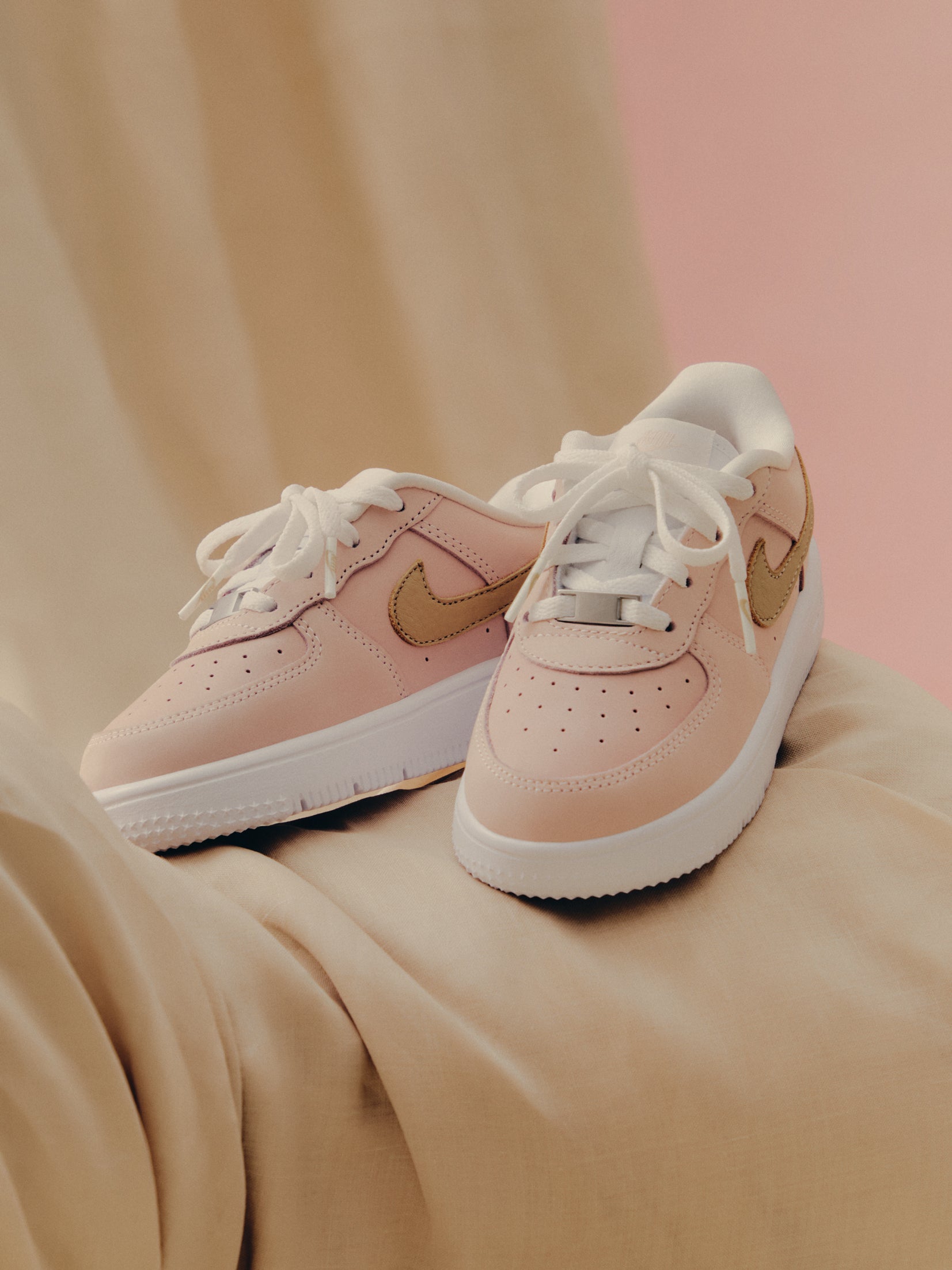 Kith x Nike Air Force 1 Low pair, pink with tan Swoosh and white sole, angled on tan fabric