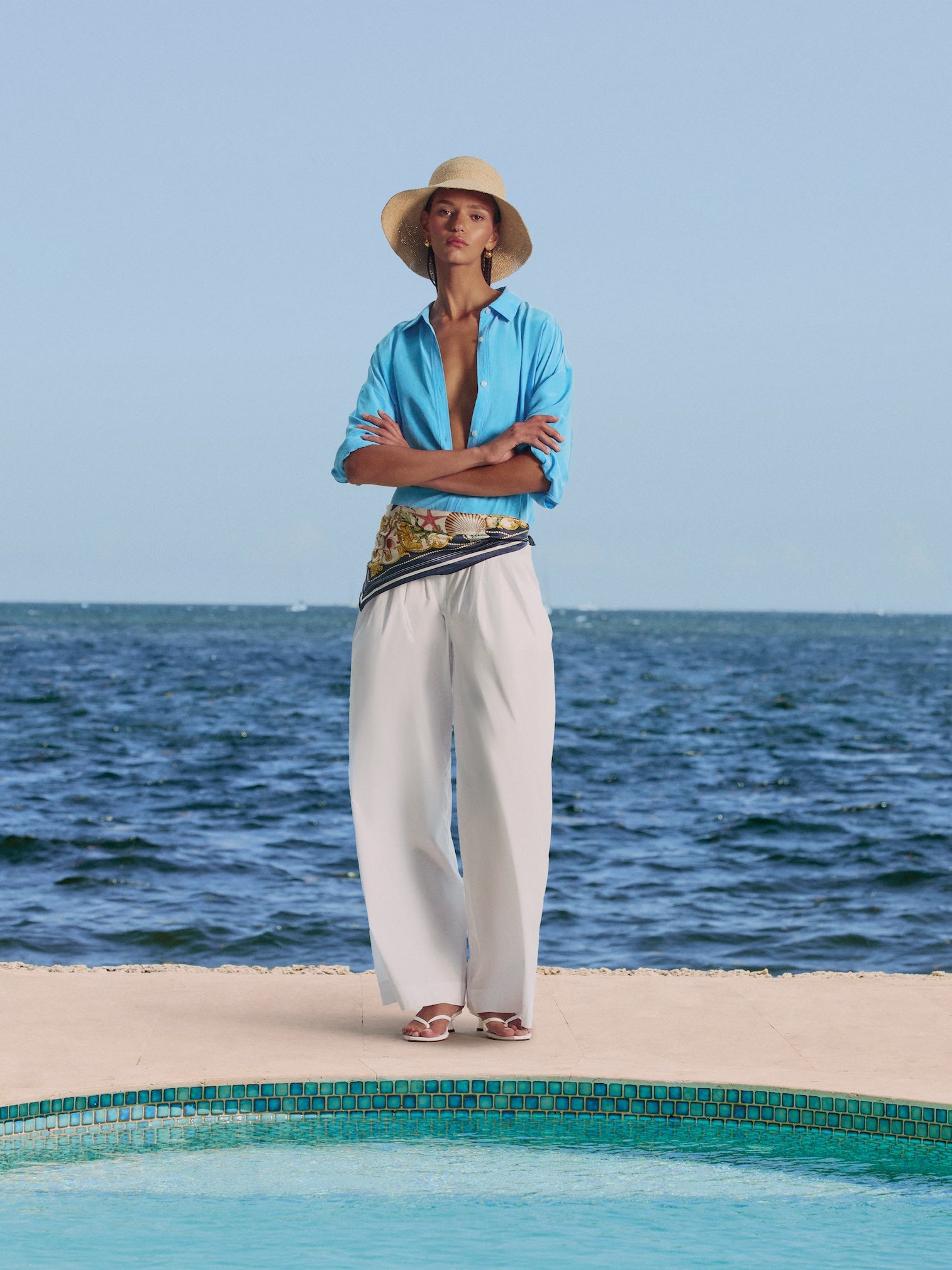 Woman poolside in a turquoise button shirt, white wide-leg trousers, straw hat and printed scarf tied at the waist