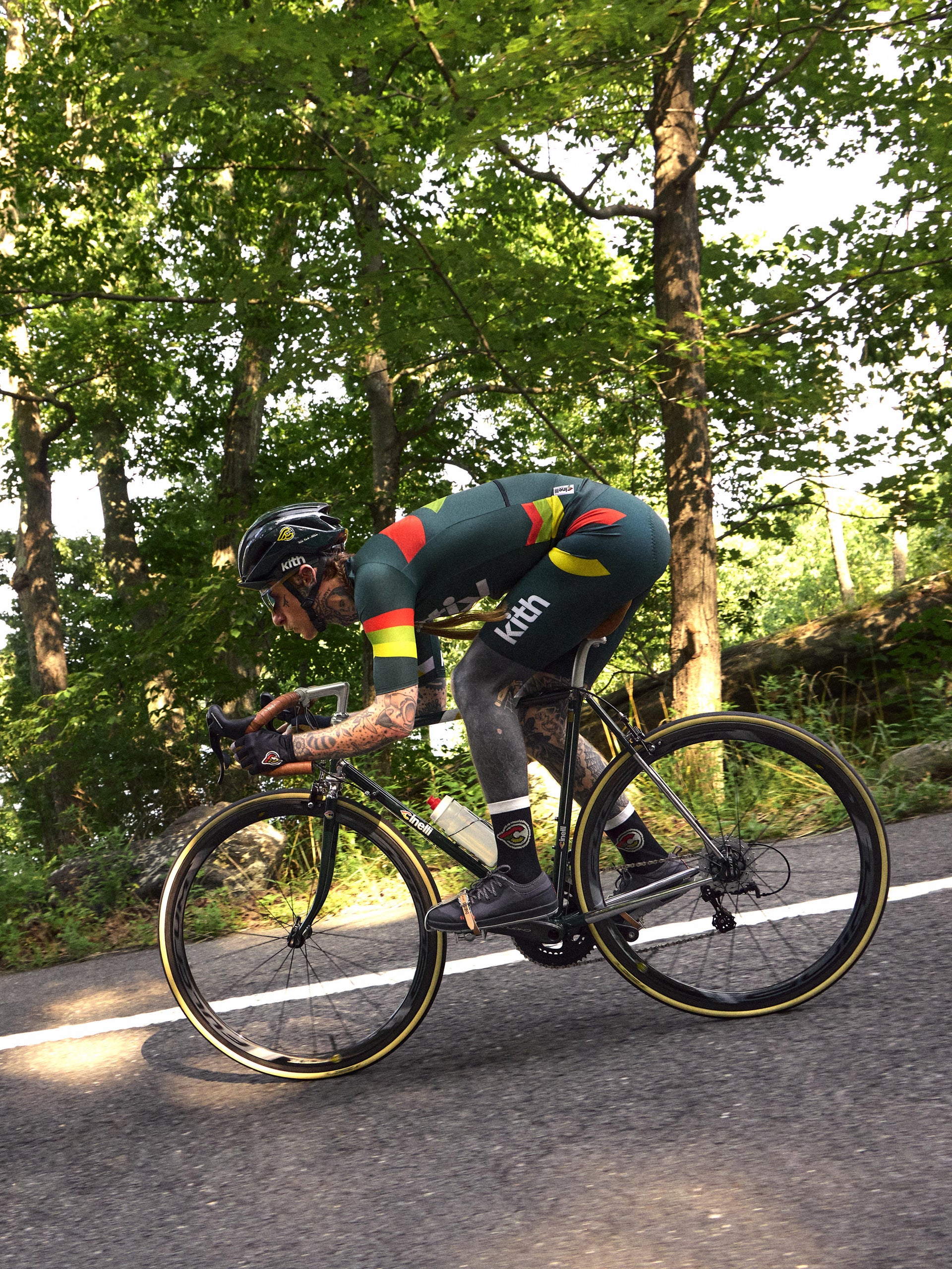 Cyclist in green Kith x Cinelli kit with red-yellow stripes riding a road bike on a tree-lined road, side view