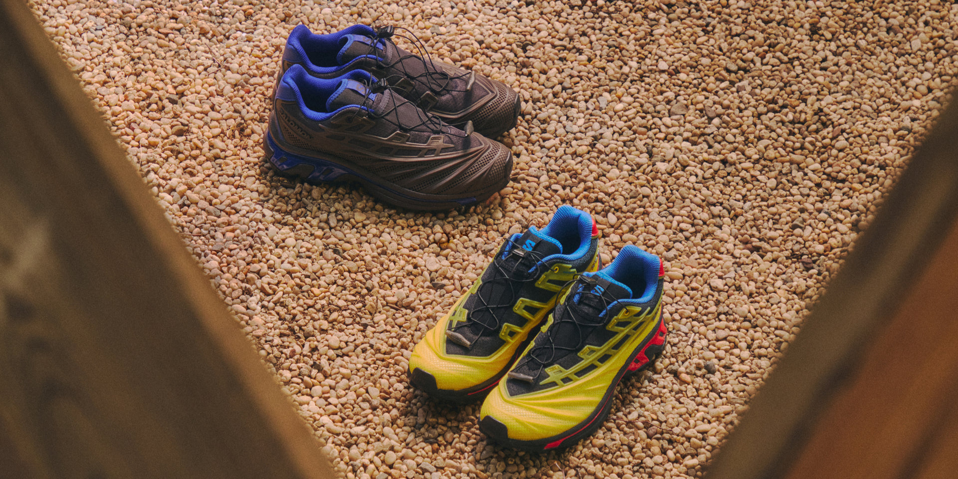 Two pairs of Salomon XT-6 sneakers on pebbled gravel, one brown and blue, one yellow, black and red, overhead view