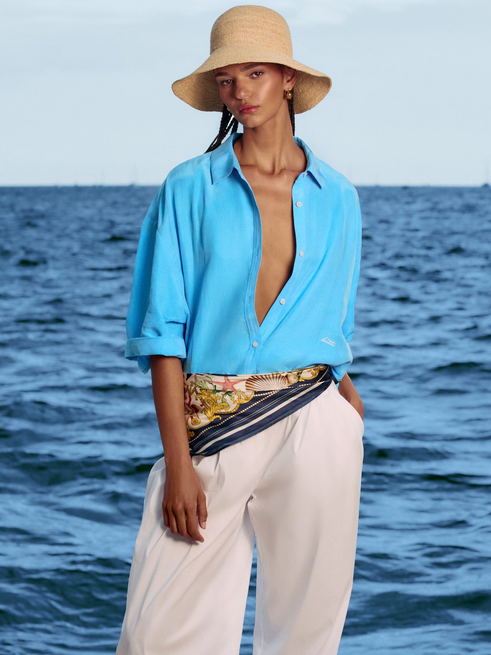 Woman by the sea in an open bright blue linen shirt, white trousers and straw bucket hat with a printed scarf at the waist