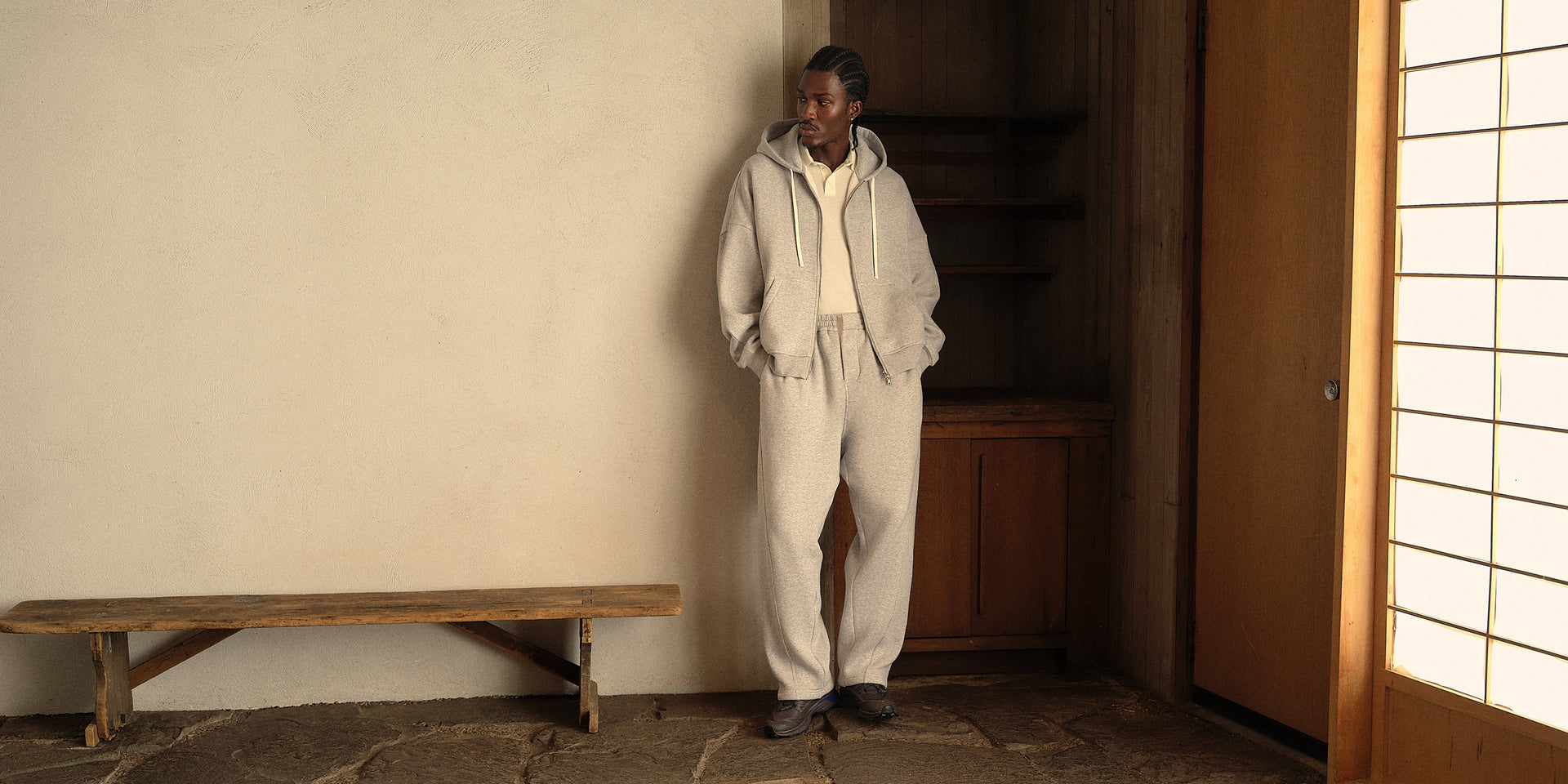 Model in grey hooded zip jacket, matching sweatpants and cream polo leaning on a wall in a sunlit room with a shoji screen
