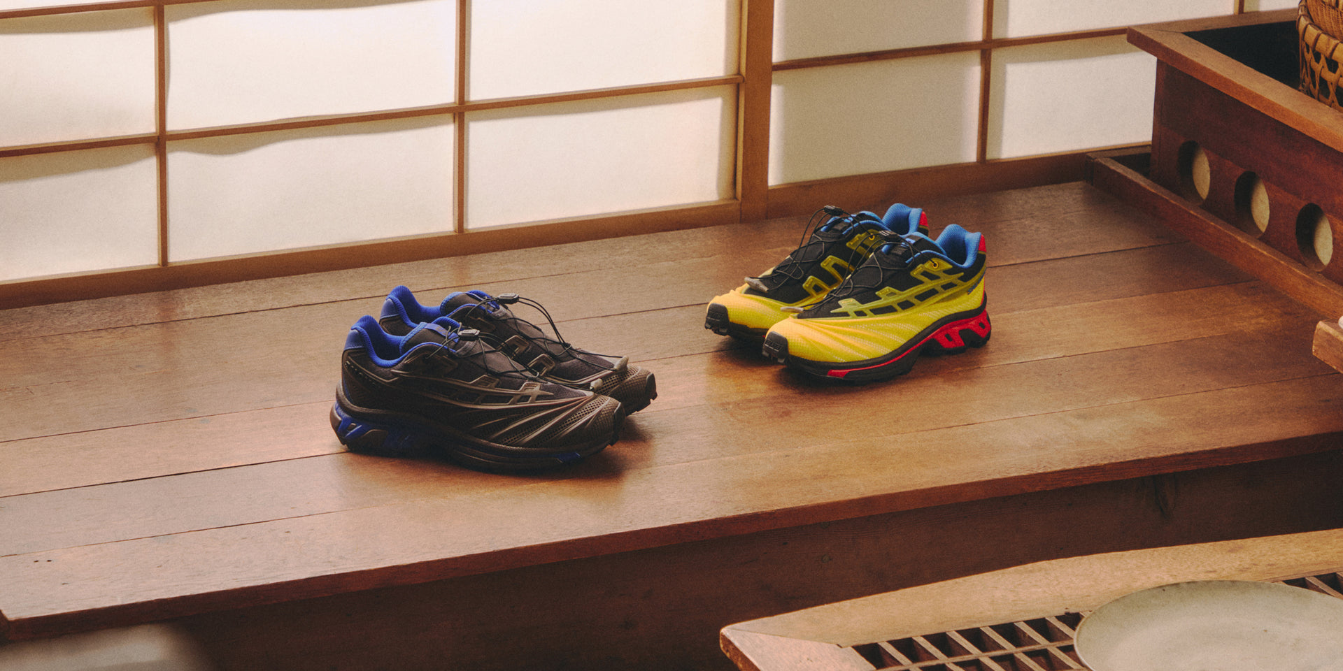 Two pairs of Salomon XT-6 sneakers on a wood ledge by a shoji screen, one black and blue, one yellow, black and red