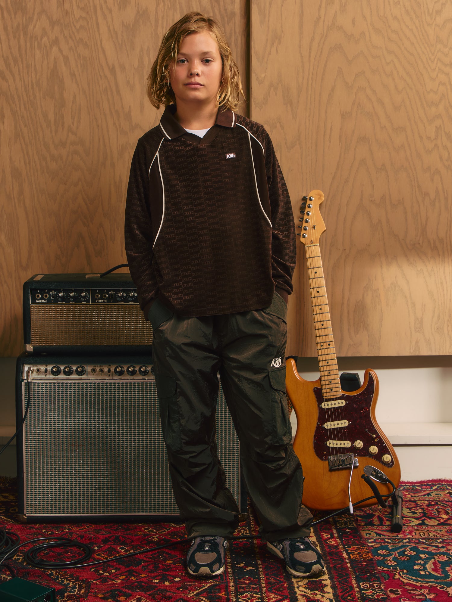 Blond boy standing between guitar amps in a brown Kith monogram long-sleeve polo and dark green nylon cargo pants with New Balance sneakers