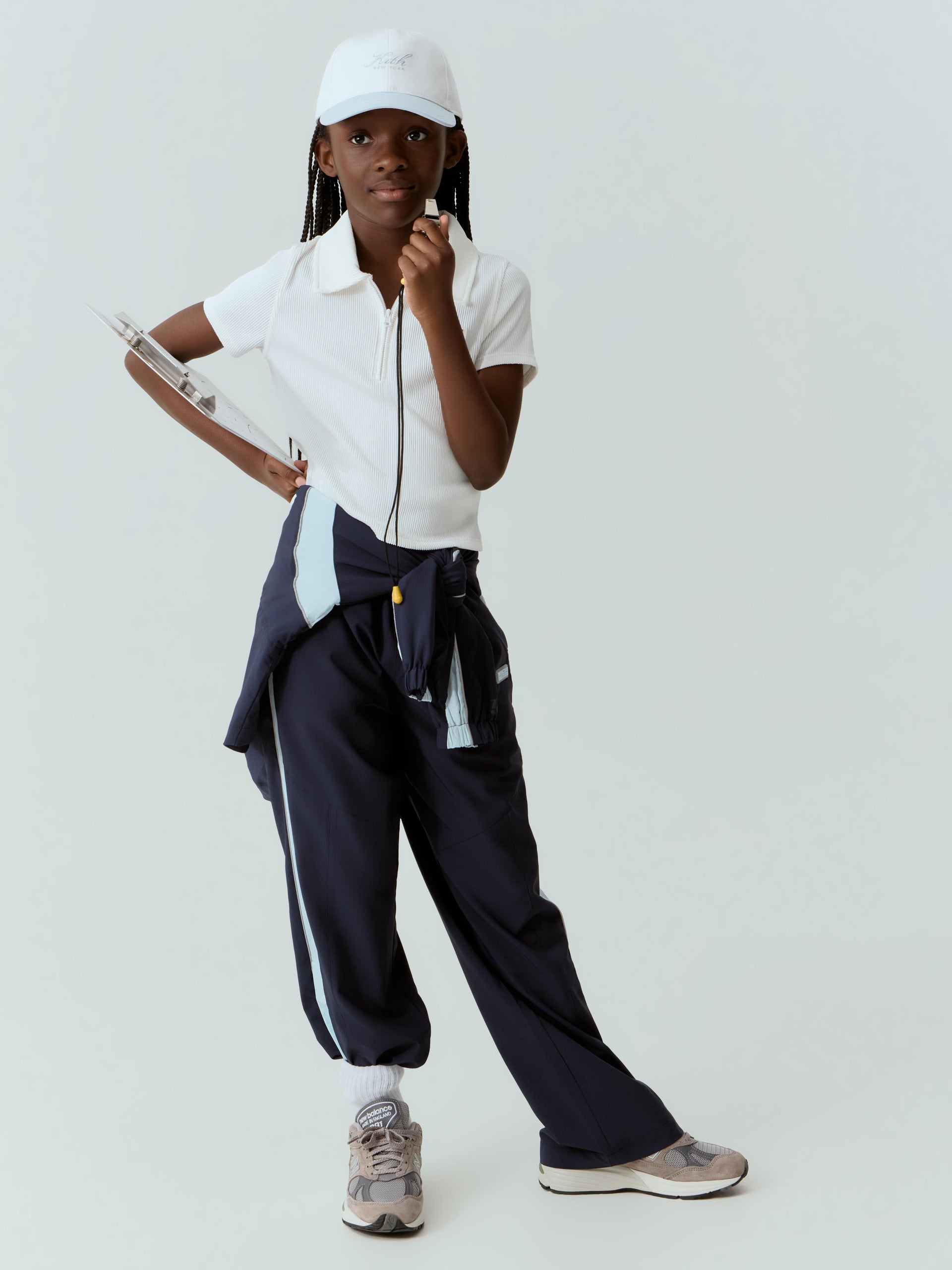 Girl in a white Kith cap and ribbed zip polo styled as a coach with whistle and clipboard, navy track pants tied at the waist, light studio
