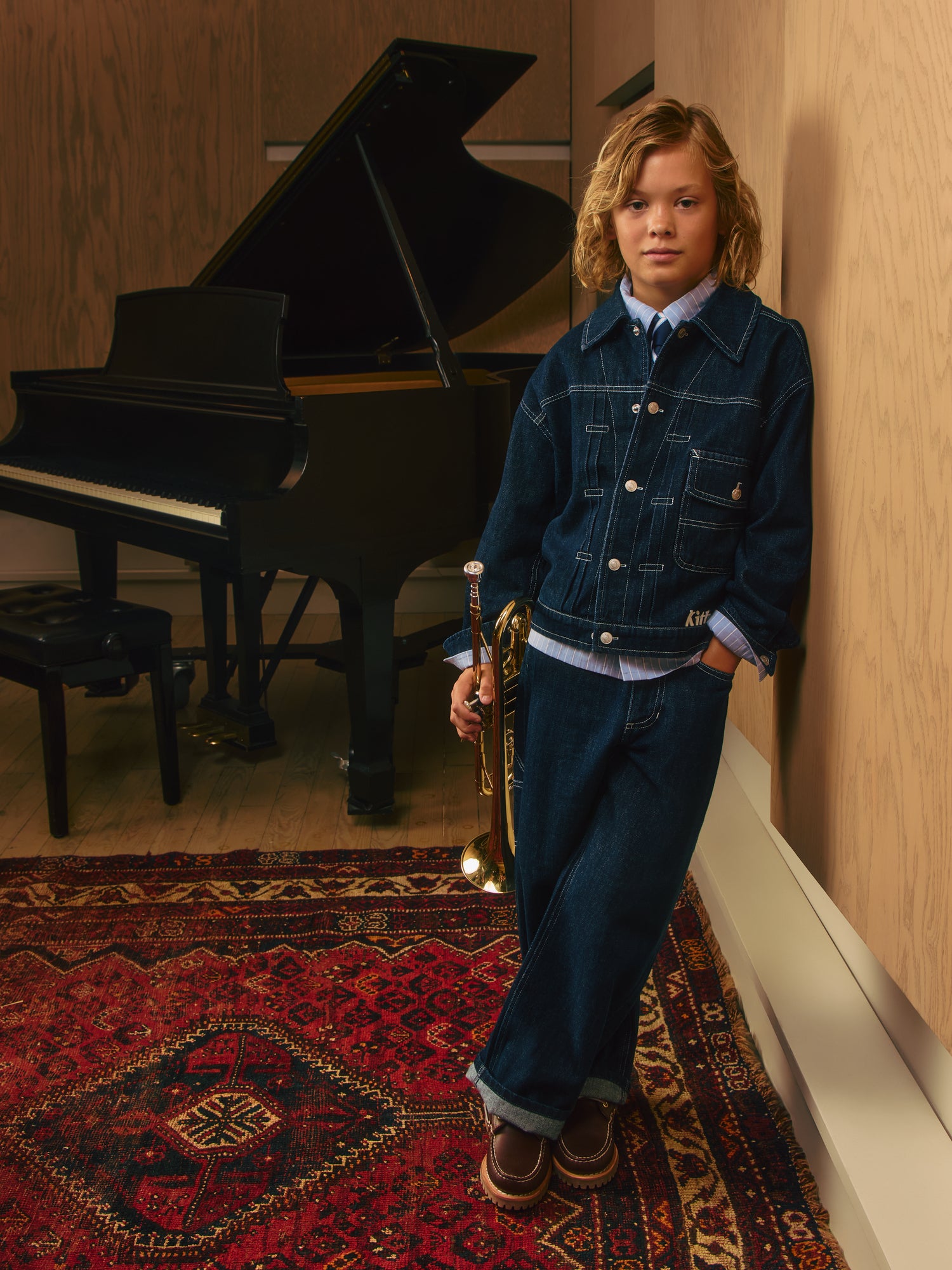 Blond boy holding a gold trumpet beside a black grand piano, wearing a dark indigo Kith denim jacket and wide-leg jeans on a red rug