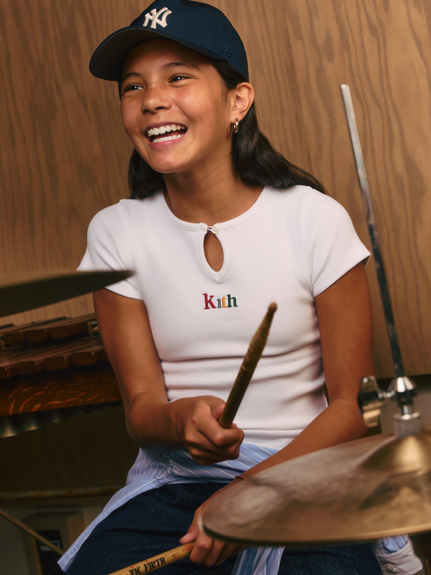 Laughing girl in a navy NY Yankees cap and white Kith ribbed keyhole tee playing drums, brass cymbal in the foreground