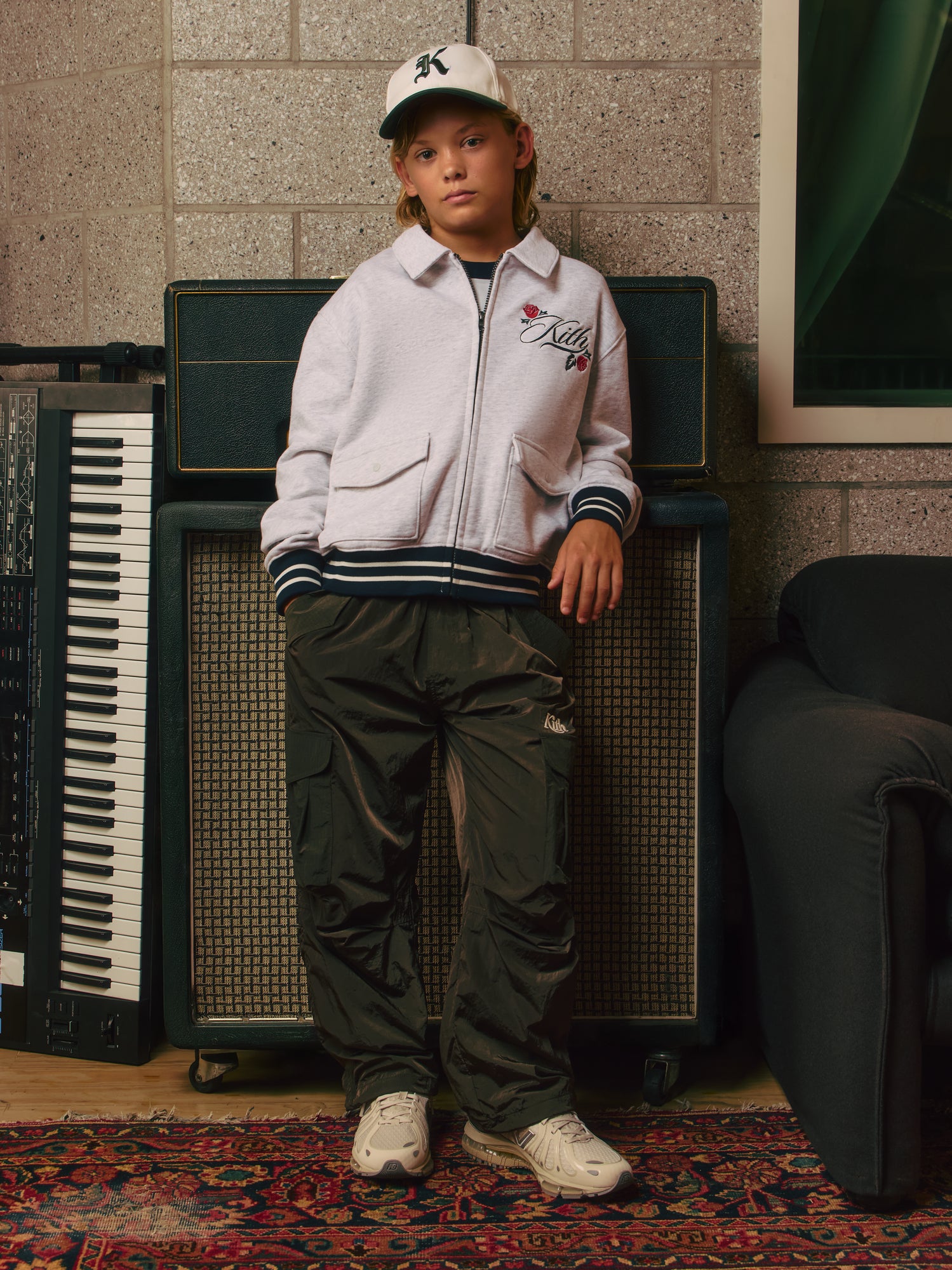 Blond boy leaning on stacked amps and a synth in a white Kith cap and grey rose-embroidered zip sweatshirt with olive cargo pants