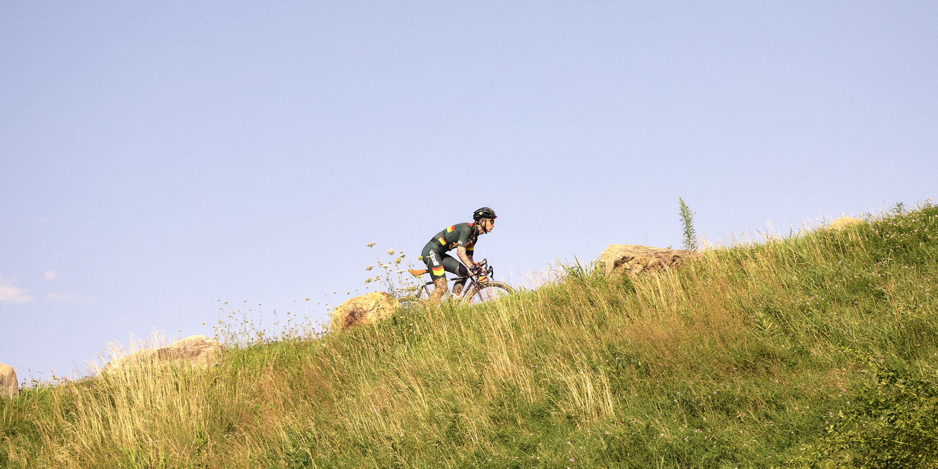 Cyclist in green Kith x Cinelli kit riding a road bike up a grassy hillside against a clear blue sky, wide shot
