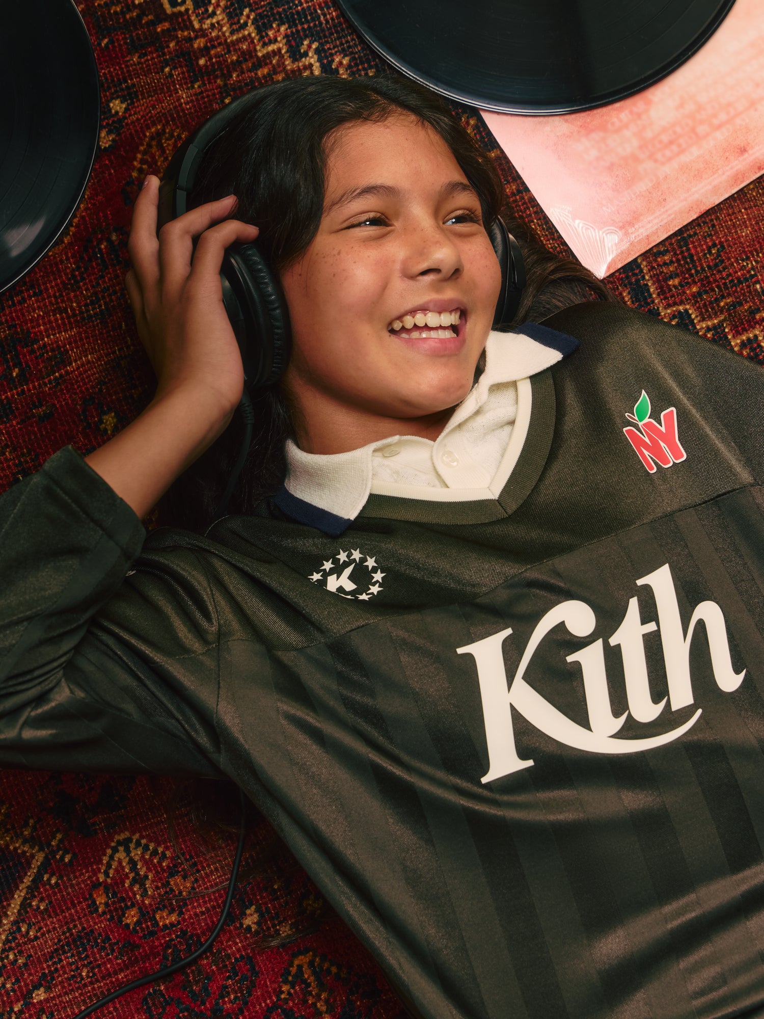 Smiling girl lying on a red rug wearing headphones and an olive Kith New York soccer jersey, vinyl records scattered around her