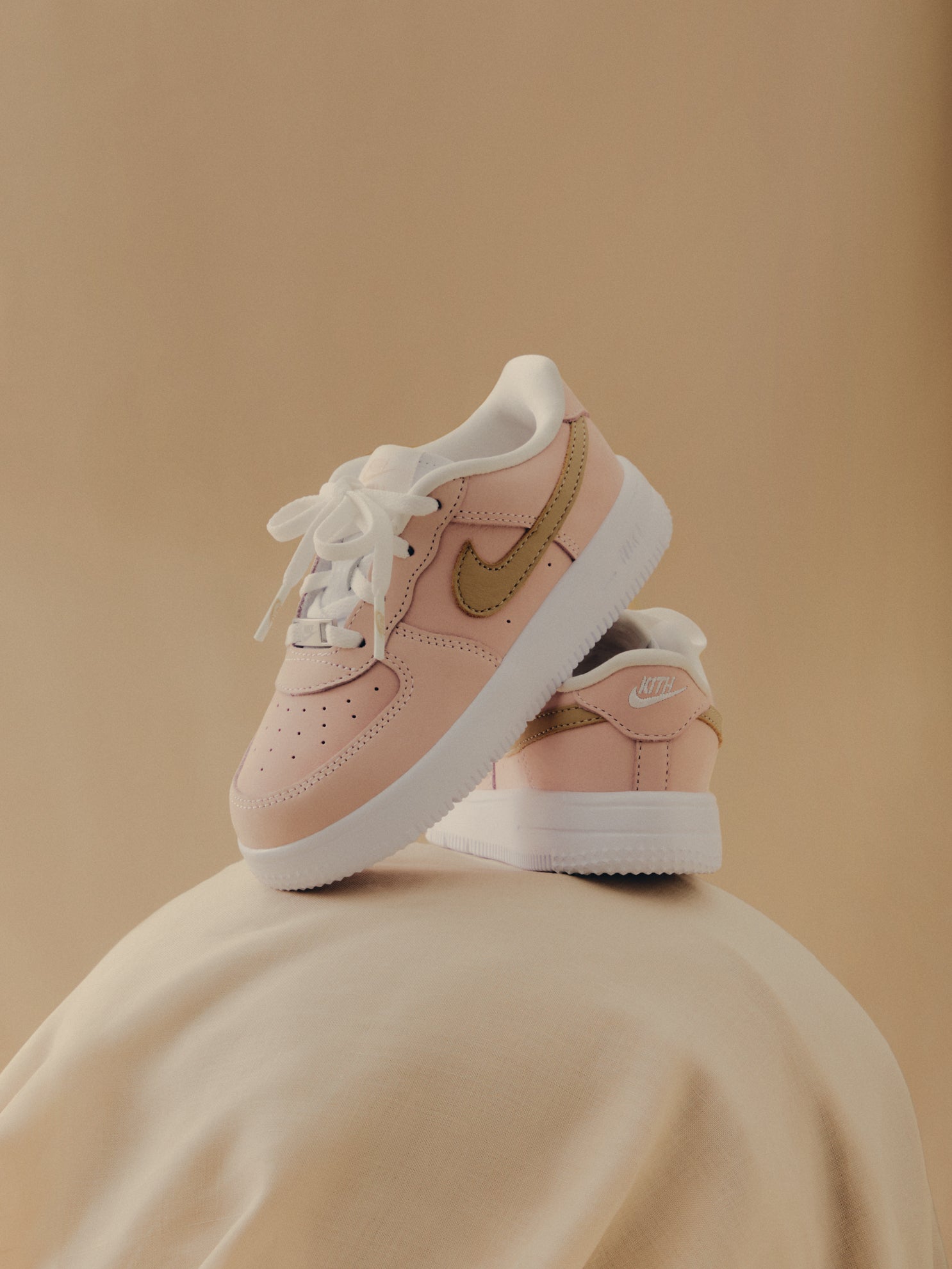 Kith x Nike Air Force 1 Low, pink with gold Swoosh and KITH heel branding, pair on cream fabric