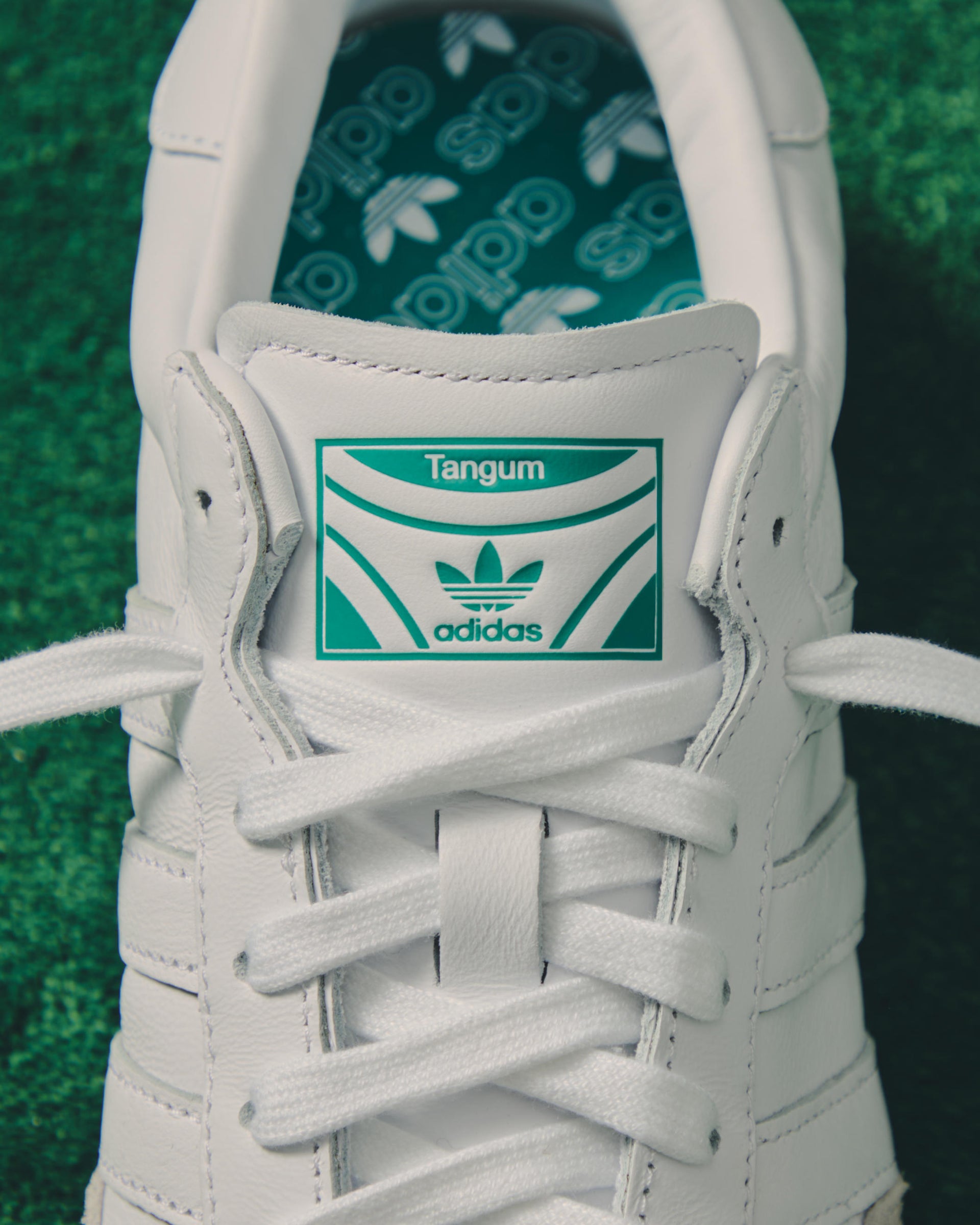 Close-up of a white leather adidas sneaker tongue with a green 'Tangum' label and Trefoil logo on green turf