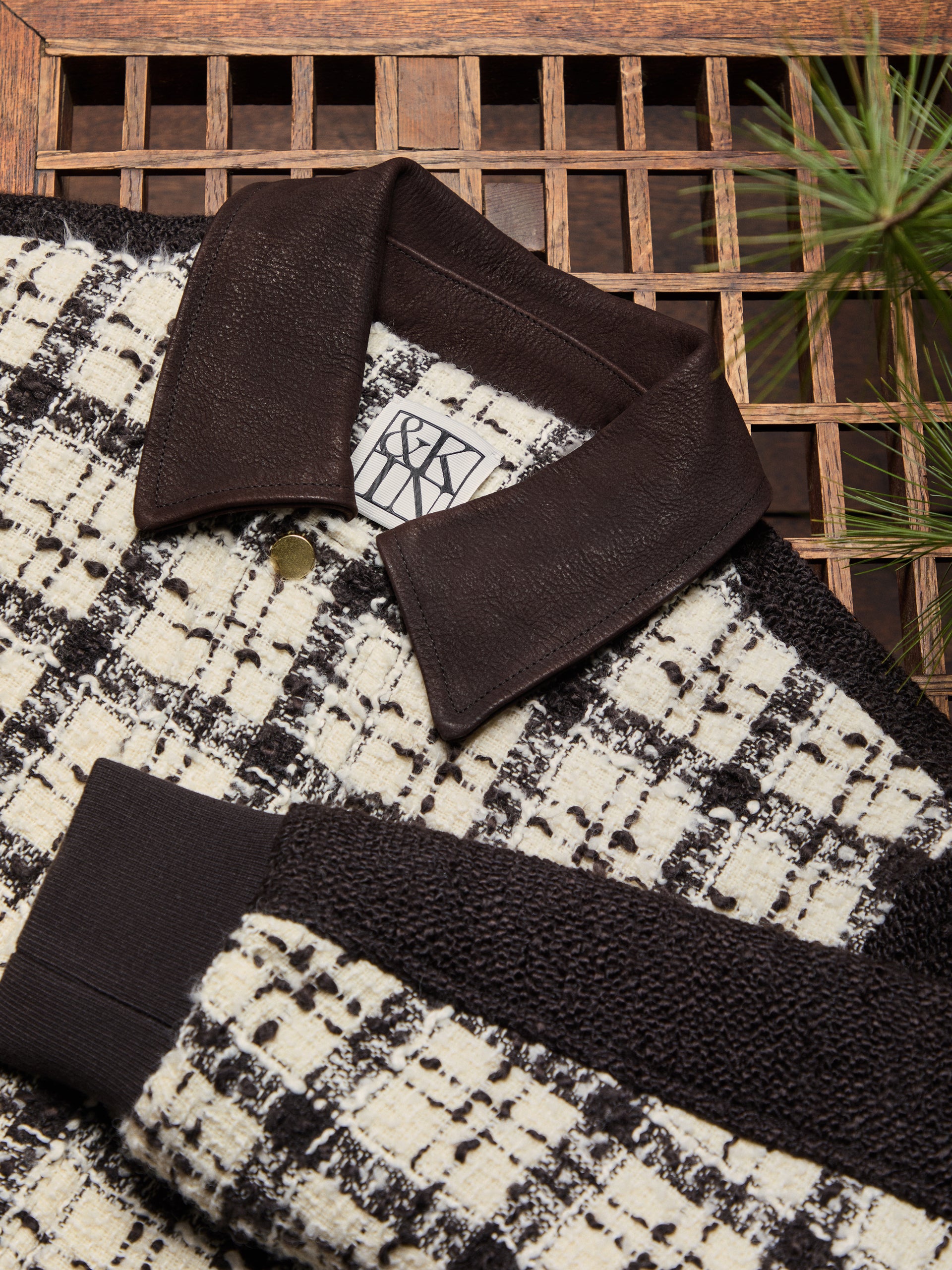 Flat-lay detail of a black-and-cream tweed jacket with dark brown leather collar, ribbed cuff and woven Kith label