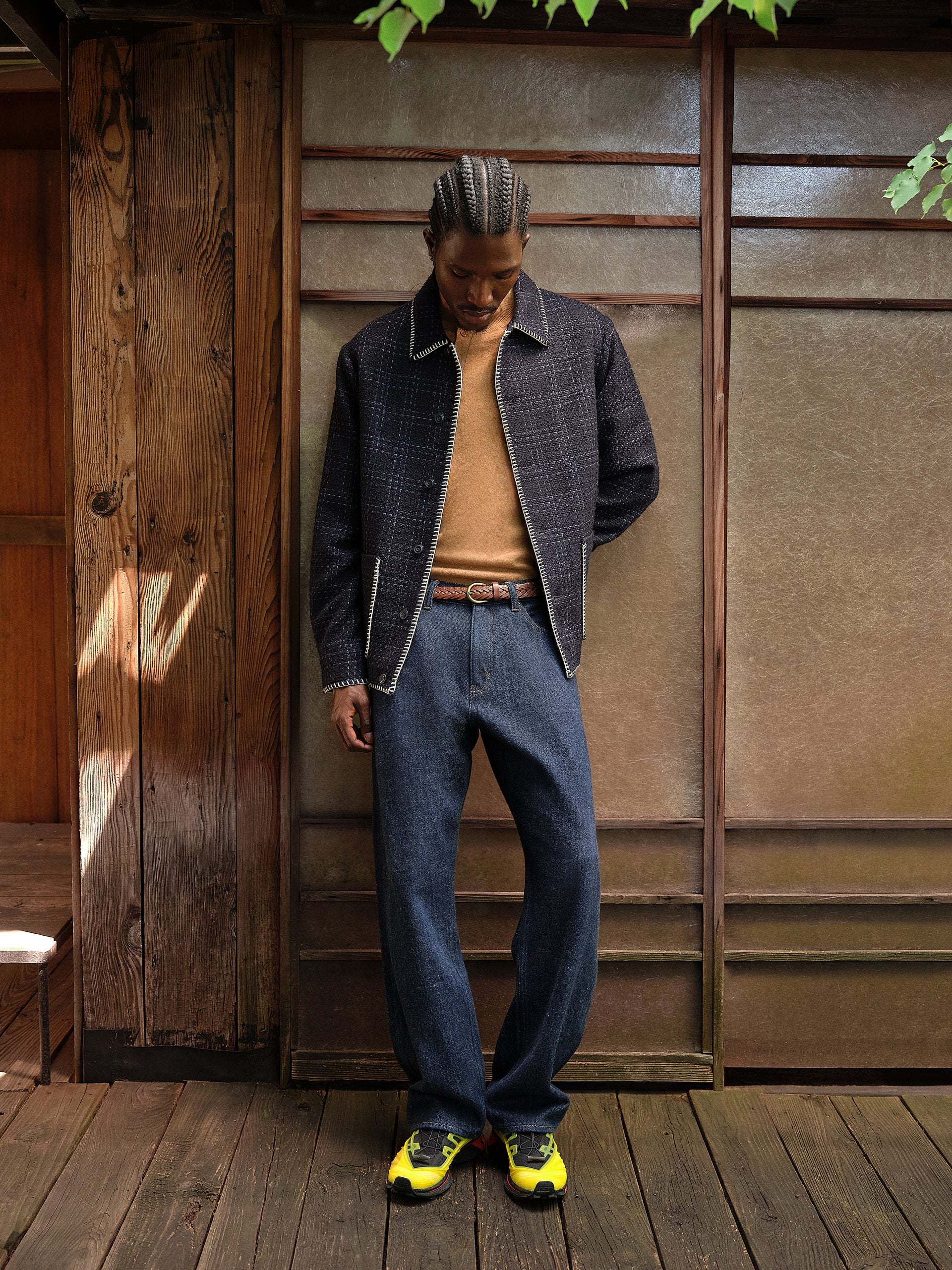 Model in a navy checked shirt jacket with blanket-stitch trim, tan knit, indigo jeans and yellow Salomon sneakers by a wall