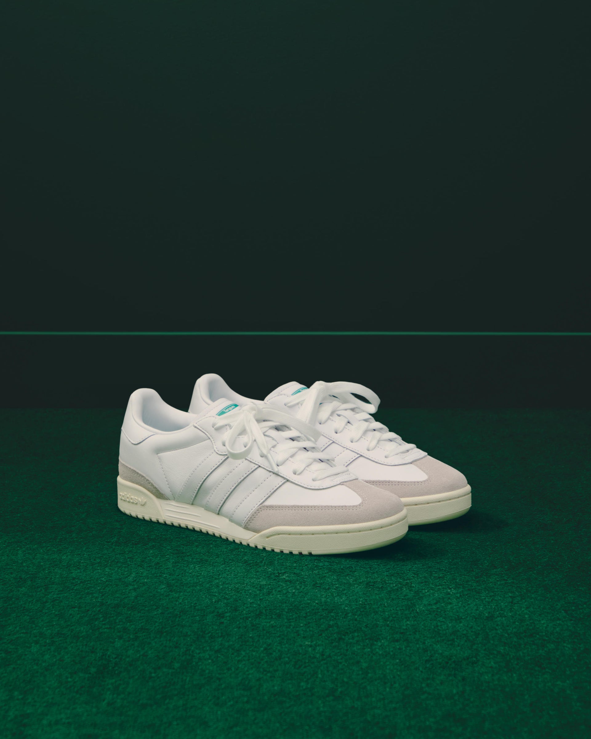 Pair of white adidas leather sneakers with green accents and cream soles on green turf against a dark green backdrop