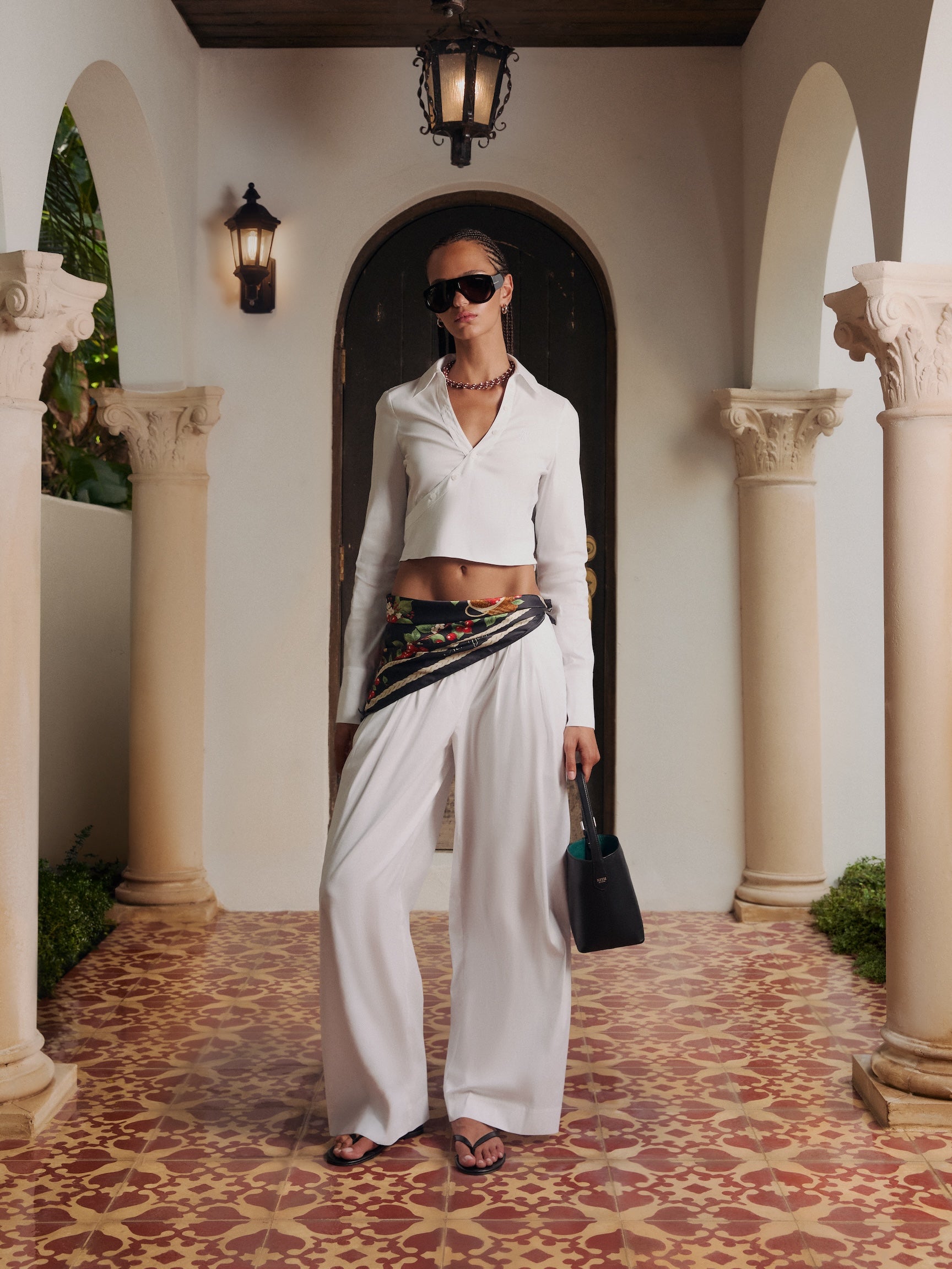 Woman in a white cropped shirt and wide-leg trousers with a floral scarf belt and black KITH bag in a tiled villa corridor