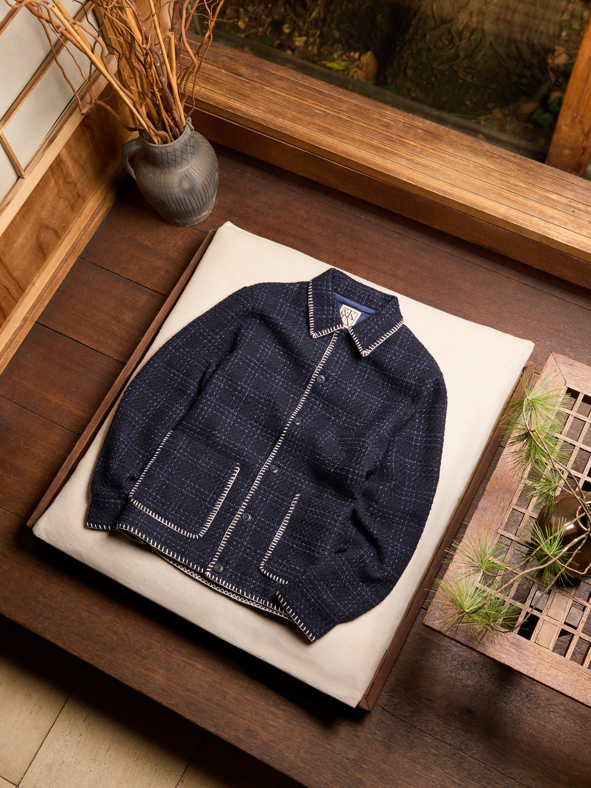Kith navy boucle plaid overshirt with cream blanket-stitch trim, flat-lay on cushion in wood-paneled interior