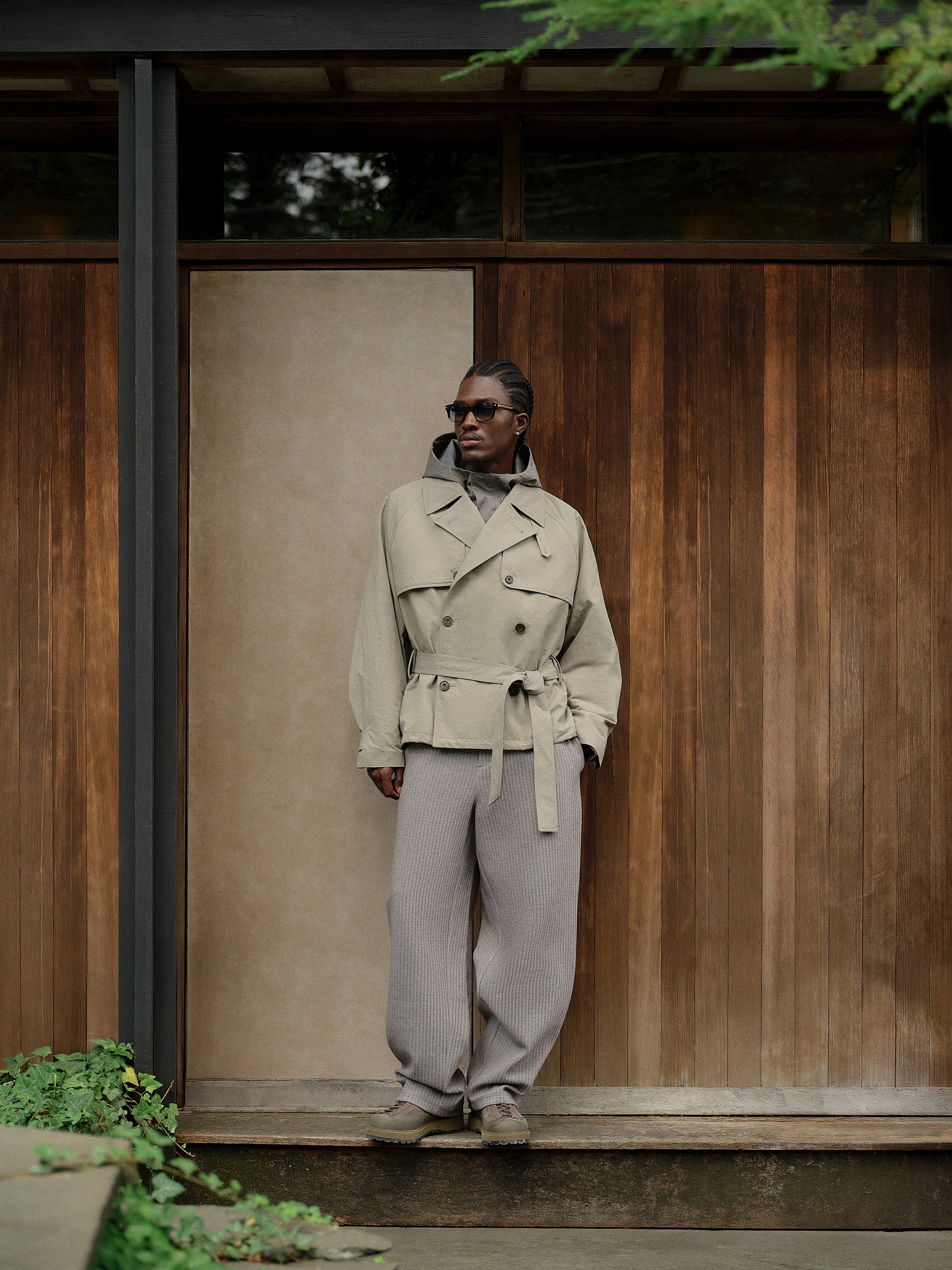 Model in beige belted short trench over grey hoodie and ribbed trousers, against wood-paneled wall, full-length