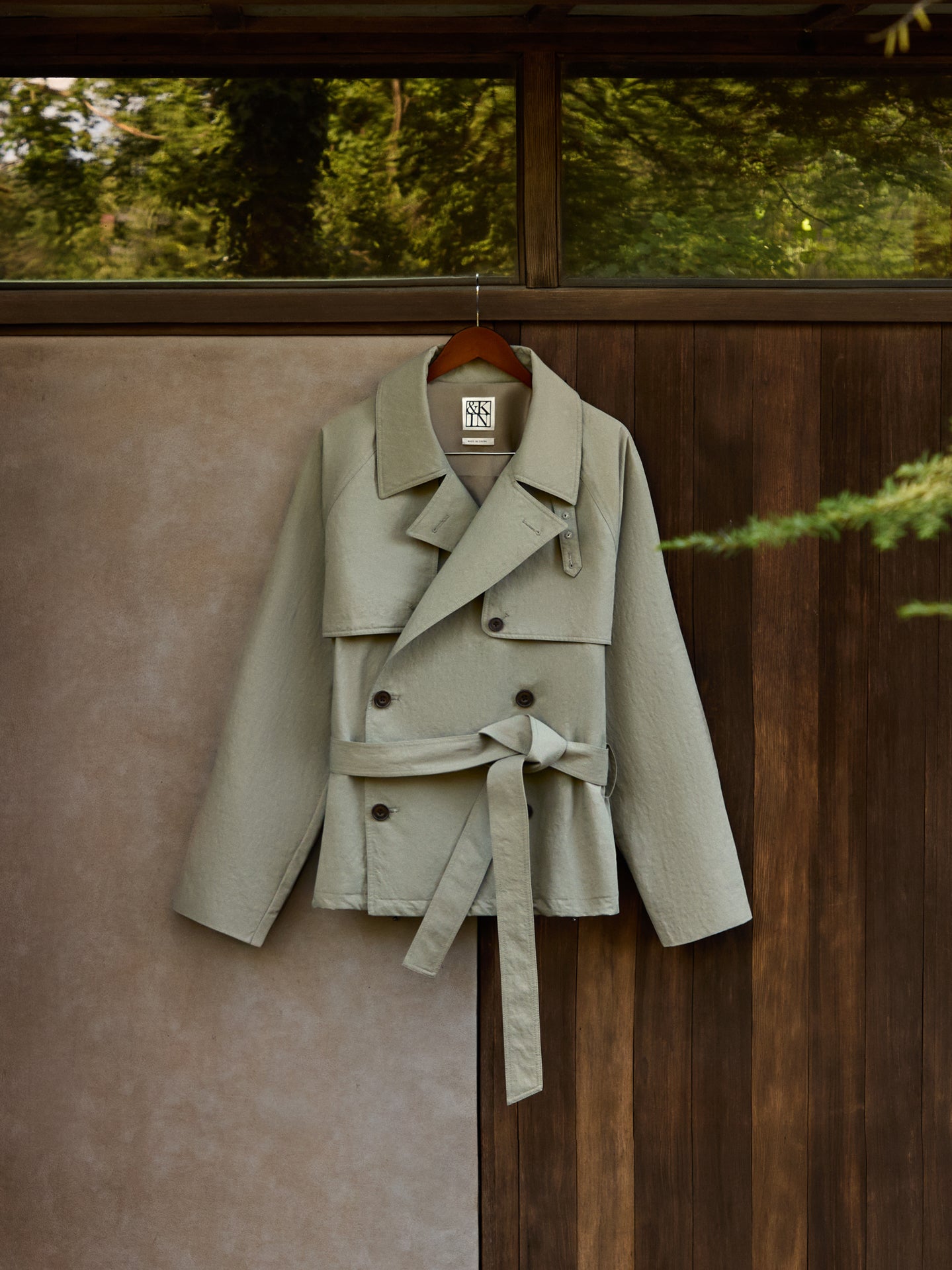 Sage green belted double-breasted short trench coat on wooden hanger against concrete and wood wall, front view