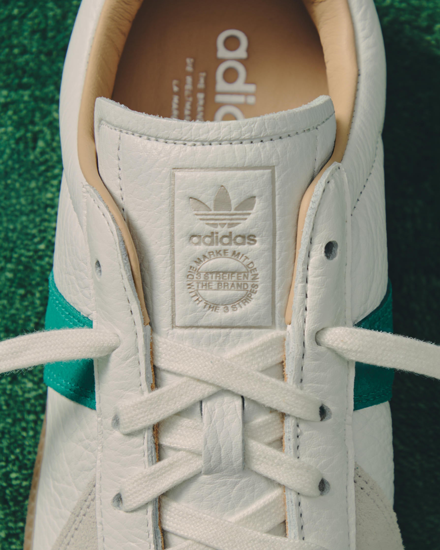 Close-up of a white leather Adidas sneaker tongue with green suede trim and tan lining, showing the debossed Trefoil logo on green turf