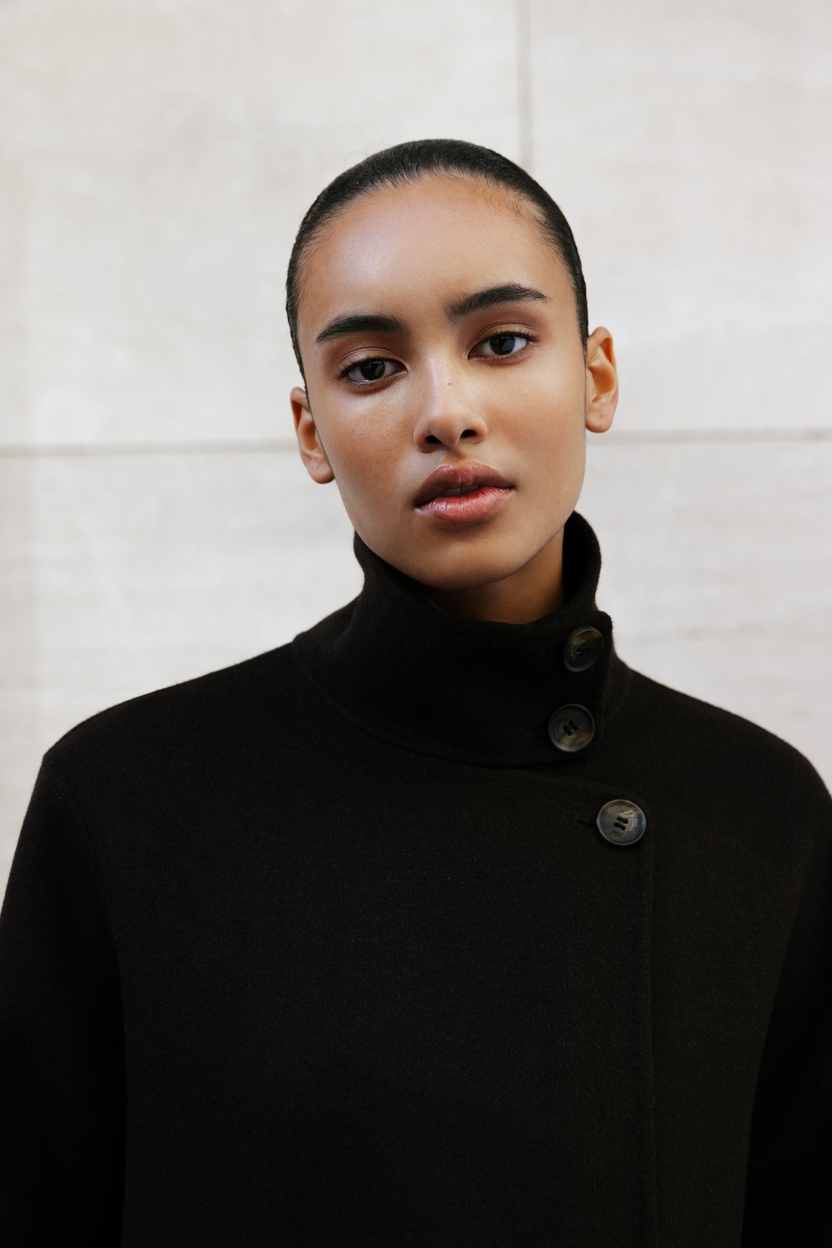 Kith woman in a black wool coat with high funnel collar and horn buttons, close portrait against pale stone wall