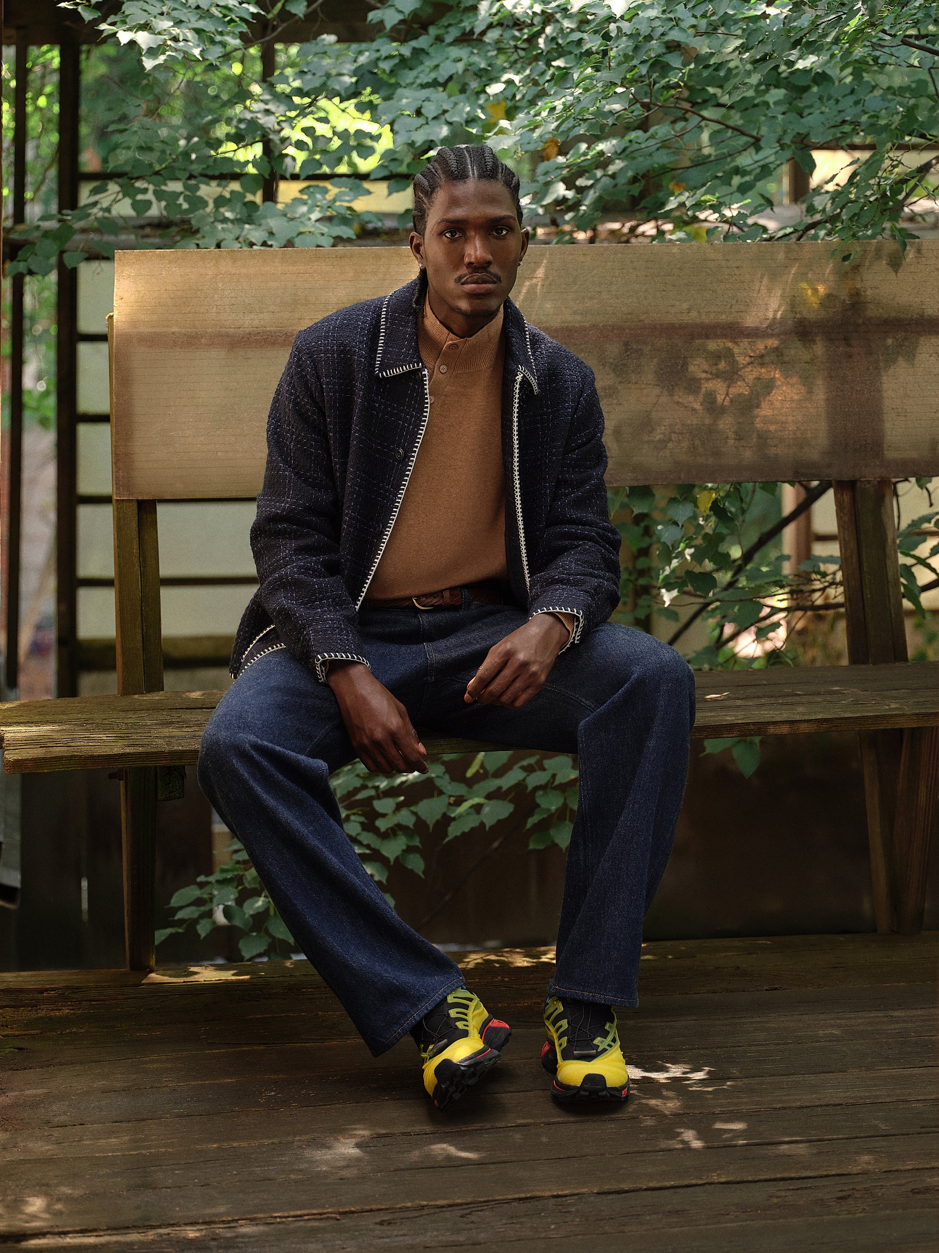 Model on wooden bench in navy blanket-stitch tweed overshirt, camel henley, indigo jeans and yellow Salomon sneakers