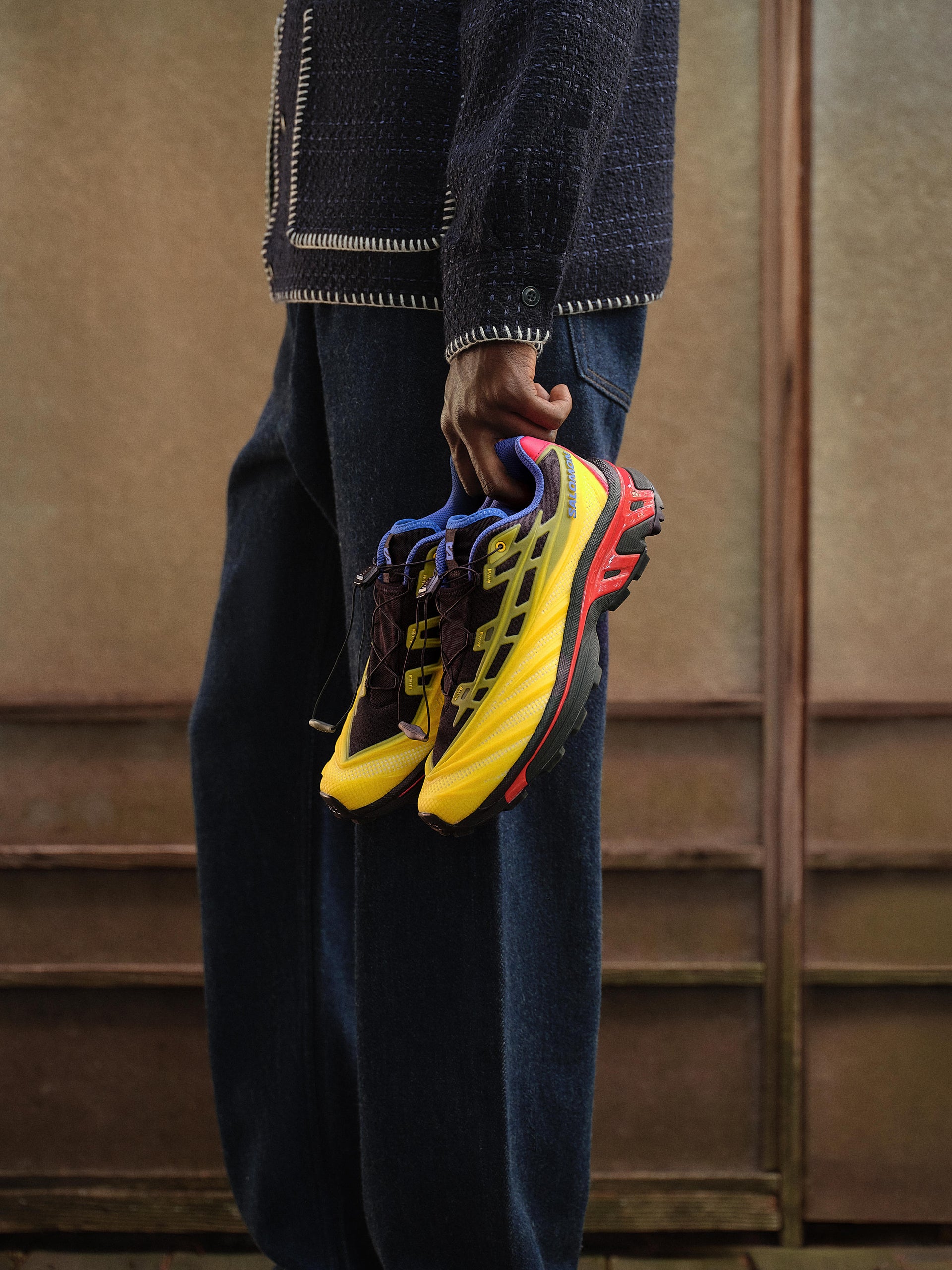 Hand holding a pair of yellow, red and blue Salomon trail sneakers beside indigo jeans, cropped detail