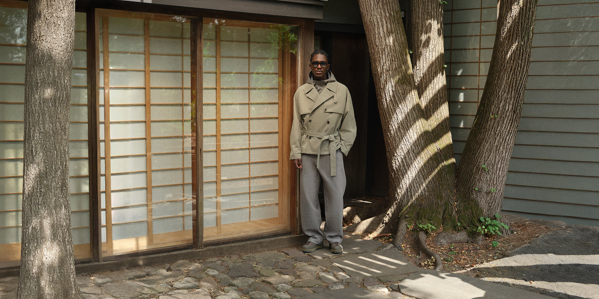 Model in olive belted trench over grey hoodie by a shoji-screen door and tree trunks, wide outdoor shot