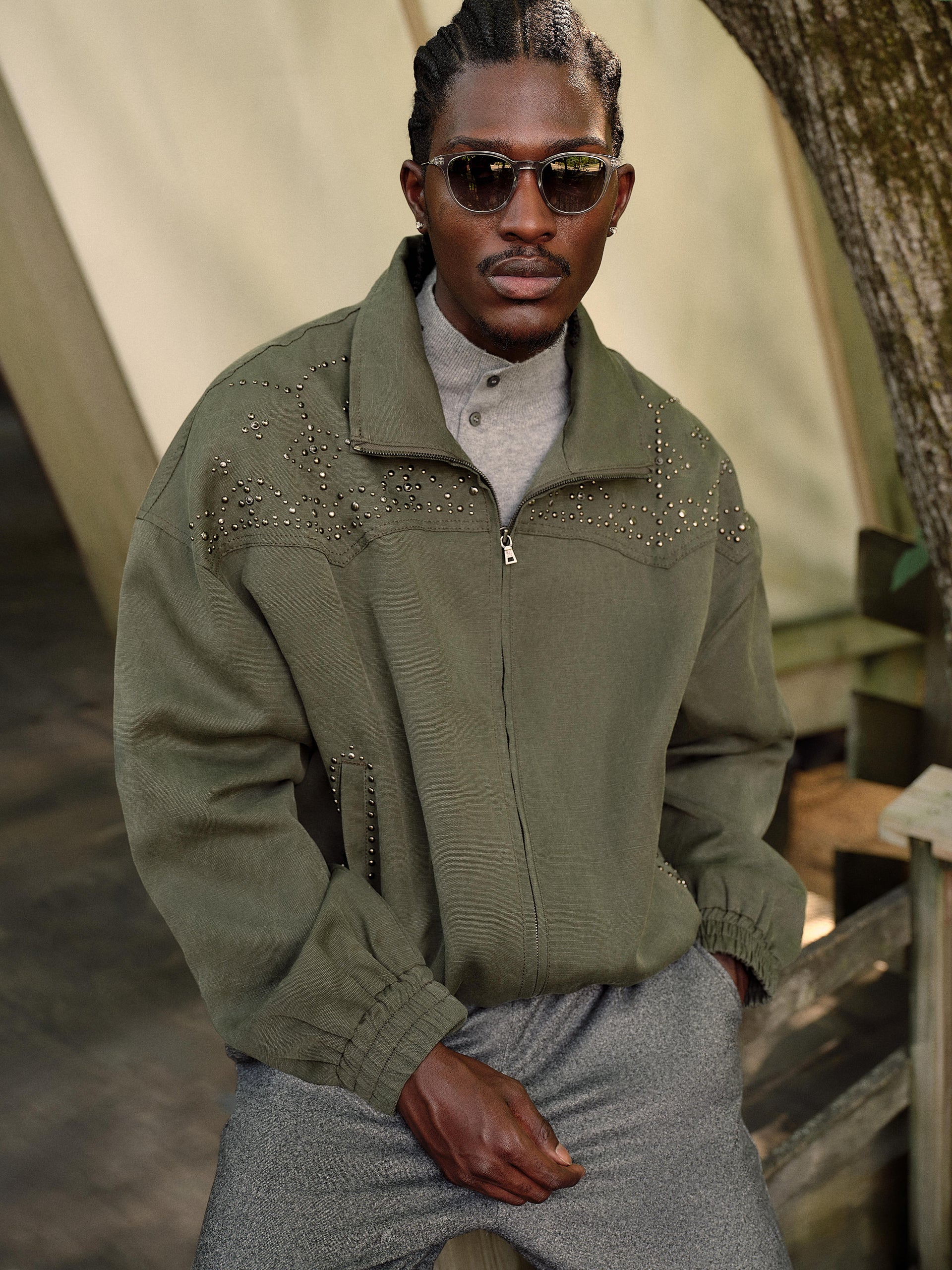 Model in sunglasses wearing olive studded zip-up jacket over grey henley, seated by a canvas tent, 3/4 view