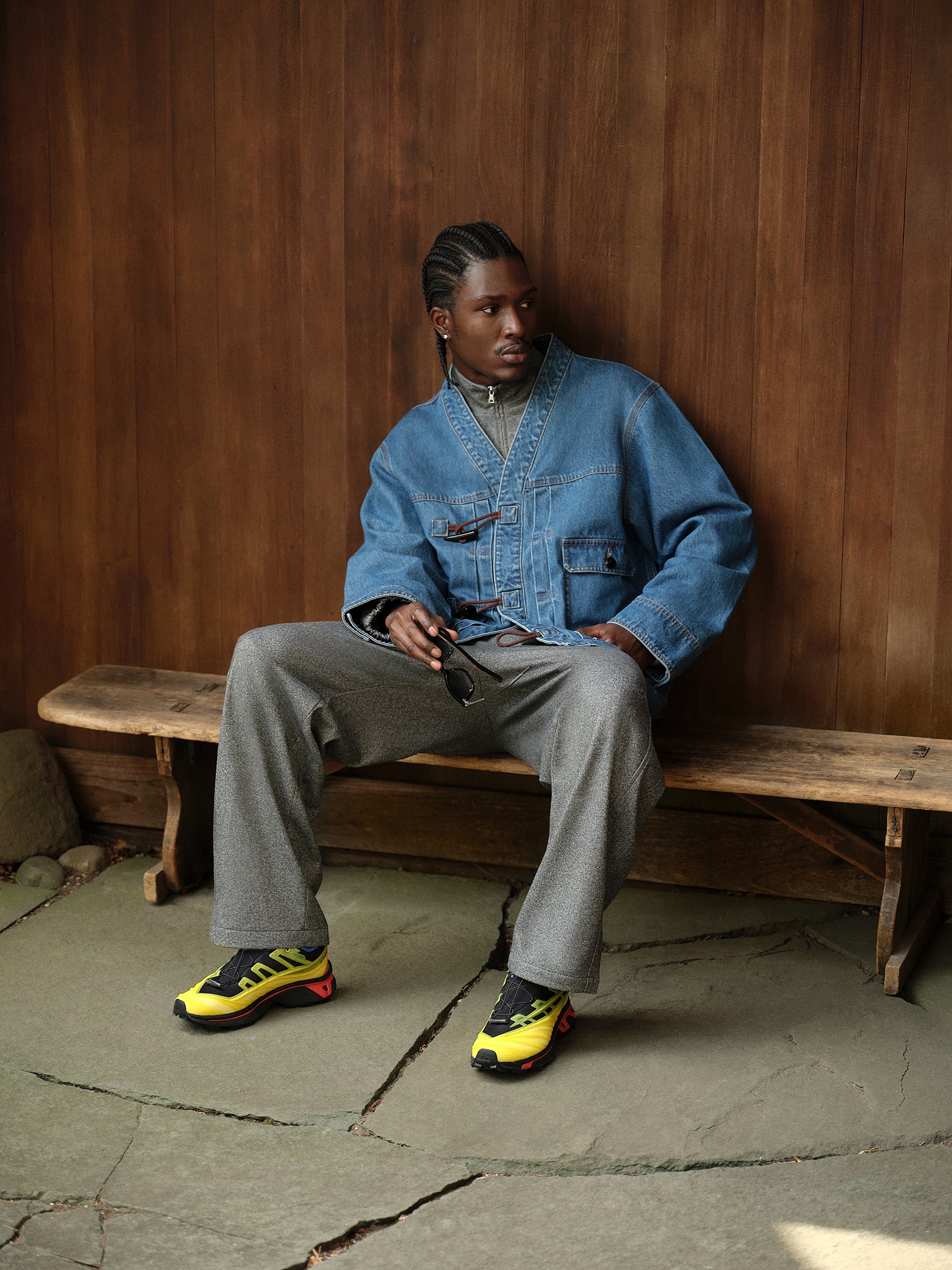 Model on bench in blue denim toggle-front haori jacket, grey trousers and yellow Salomon sneakers, holding sunglasses