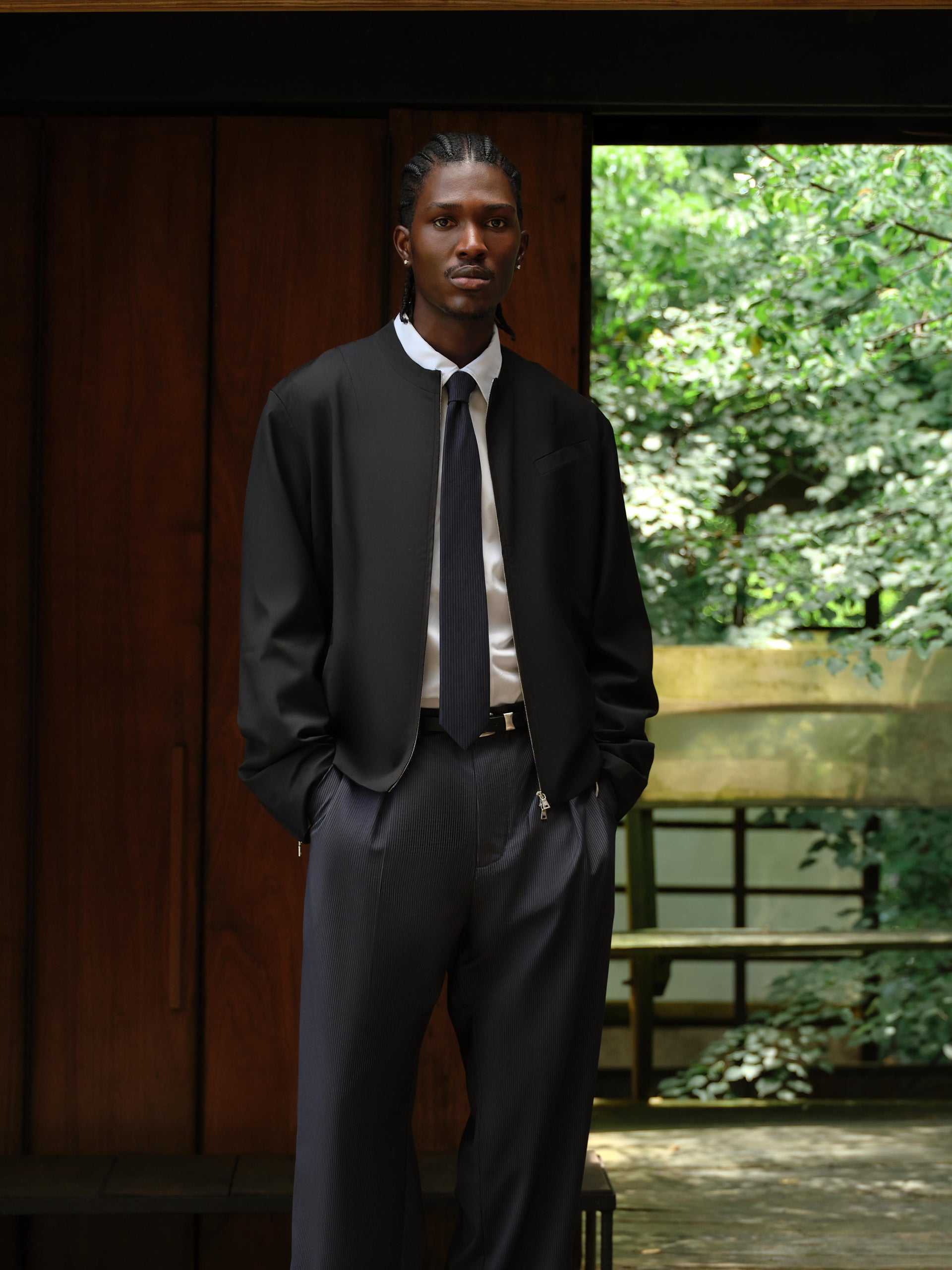Model in black zip bomber, white shirt with navy tie and navy pinstripe trousers in a wood-house doorway, full-length