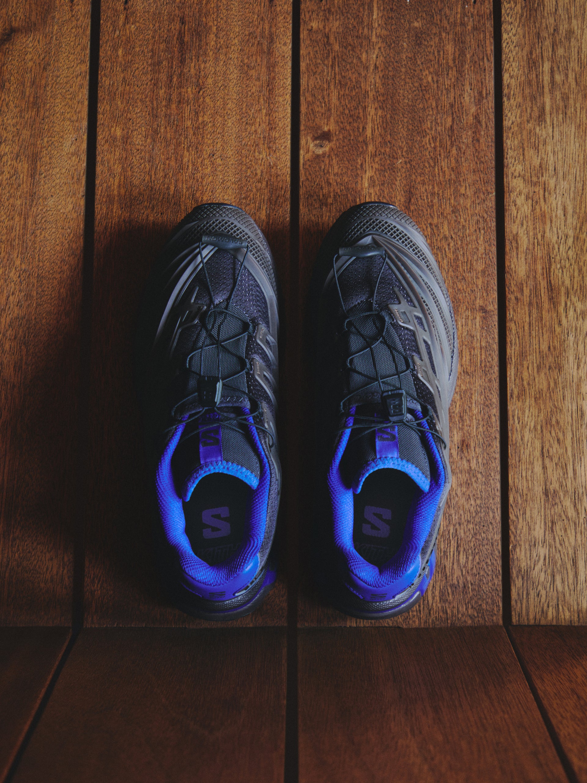 Overhead flat-lay of black and grey Salomon XT trail sneakers with blue laces on a wood-plank floor