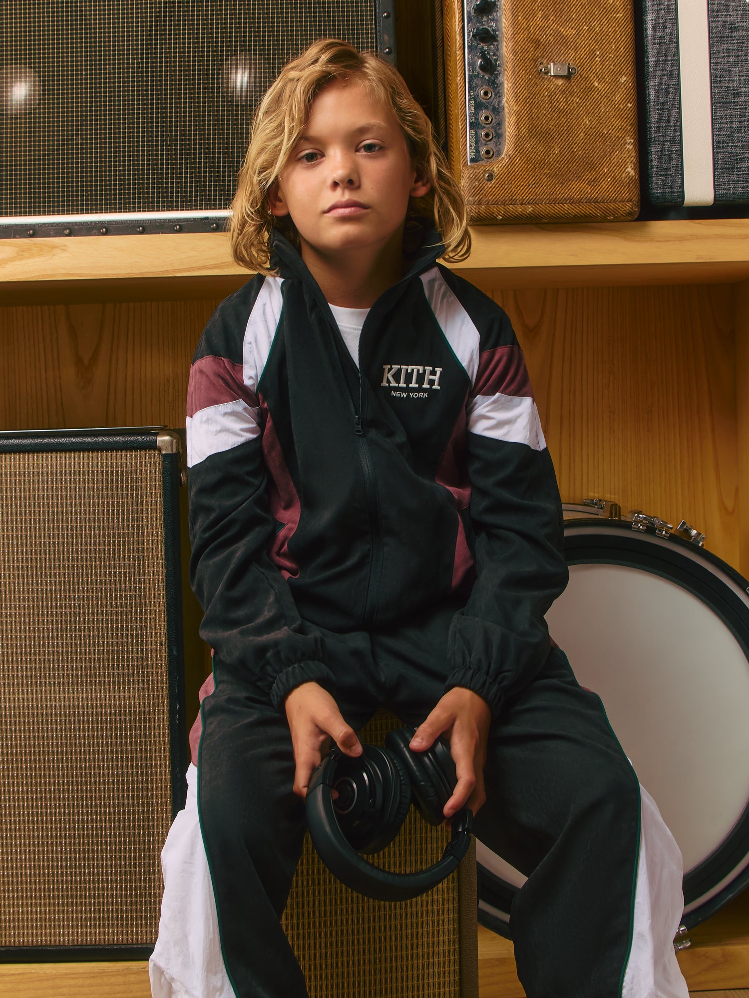 Blond boy on a vintage amp in a black Kith New York velour track jacket with maroon-and-white panels, holding black headphones