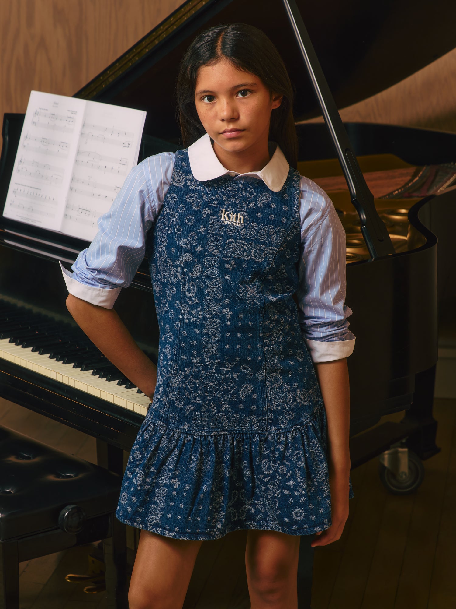 Girl in an indigo Kith New York paisley denim drop-waist dress over a blue pinstripe shirt beside a black grand piano