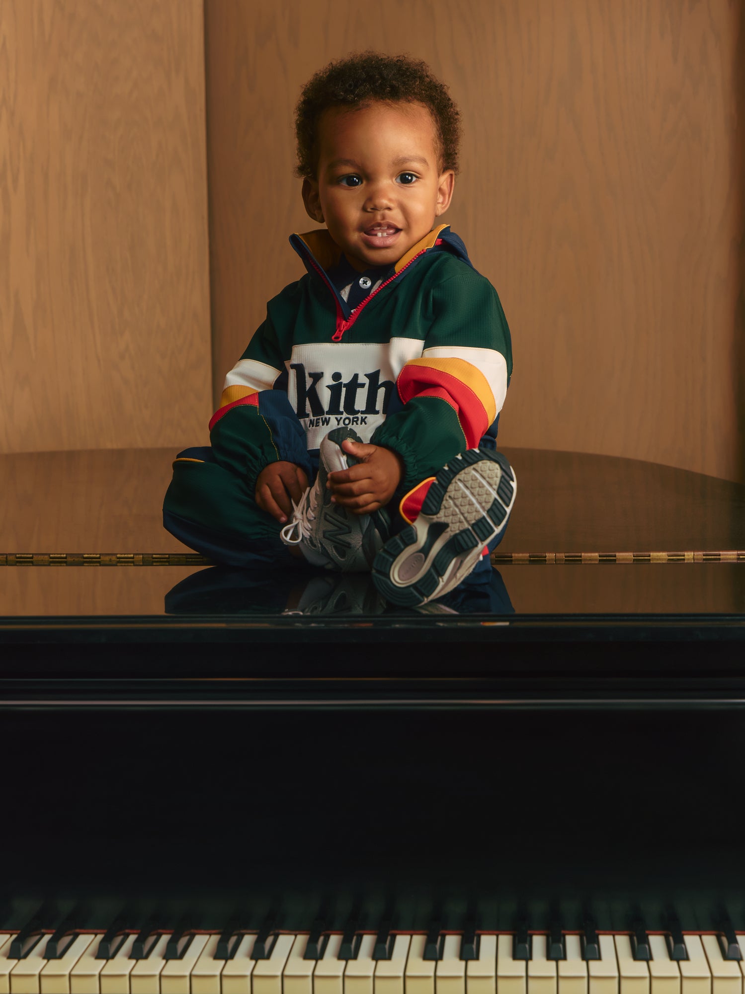 Toddler seated on a black grand piano in a green, navy and red Kith New York colorblock jacket and New Balance sneakers