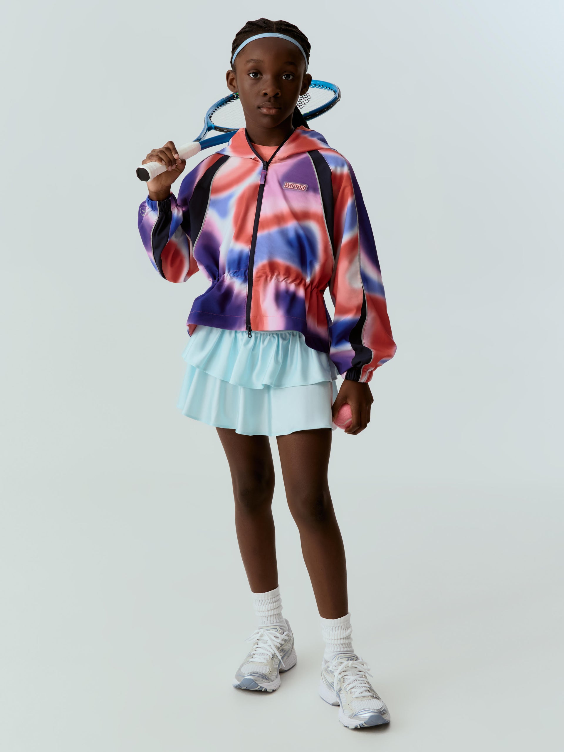 Girl tennis player in a colorful abstract-print Kith zip jacket and mint tiered skirt, racket on her shoulder, on a gray studio backdrop