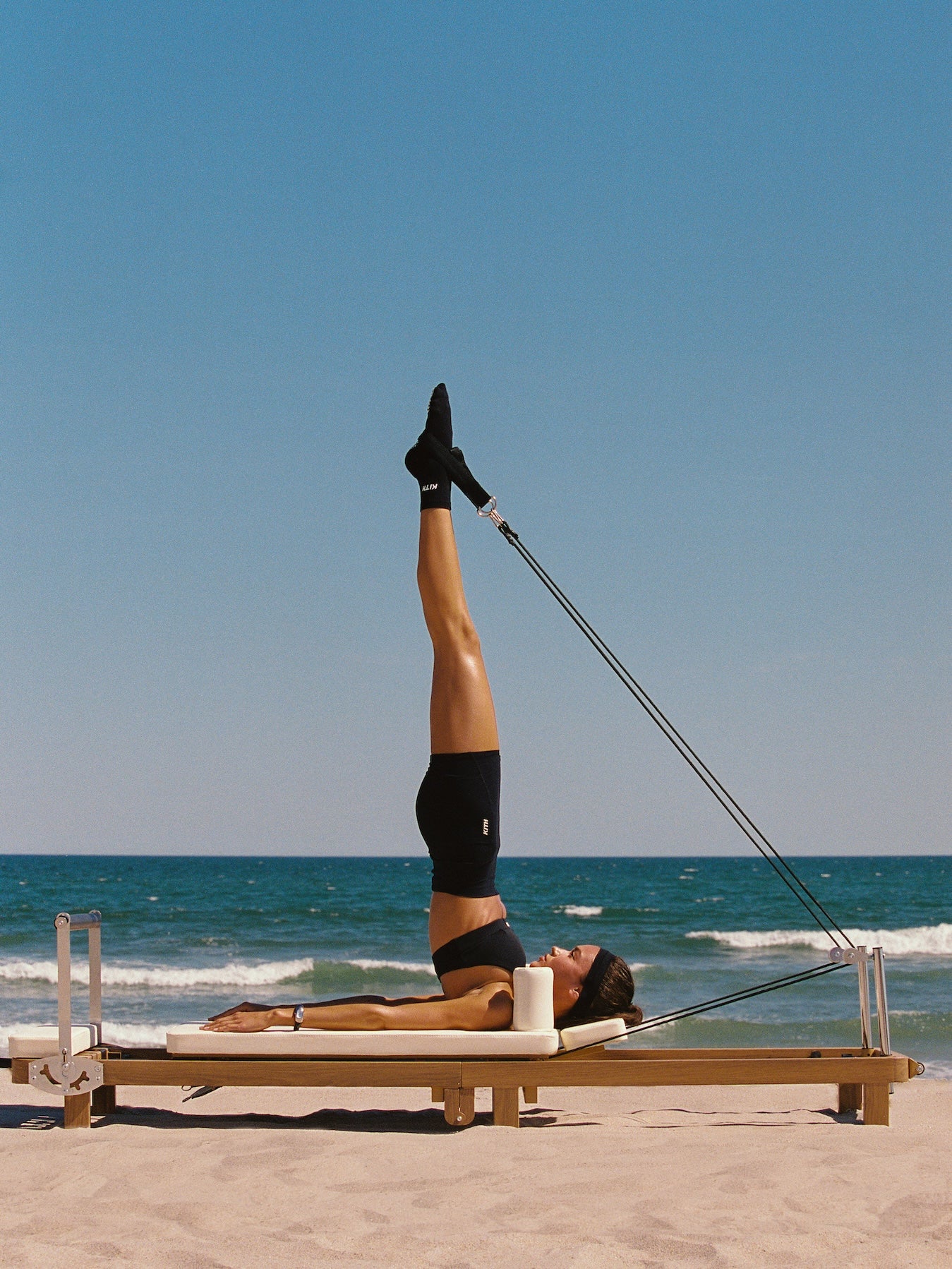 Woman on a wooden pilates reformer on the beach in a black KITH sports bra and shorts with legs raised toward a blue sky