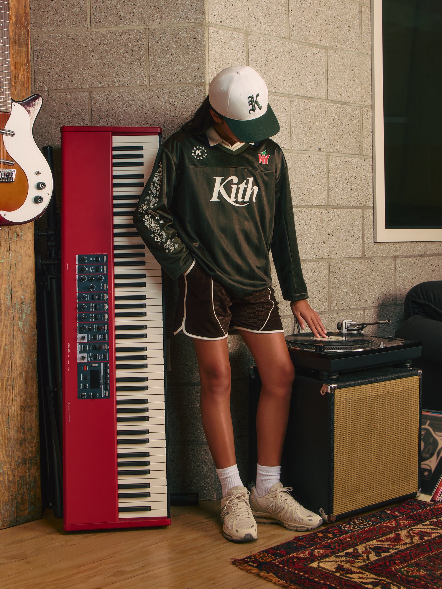 Girl in olive Kith NY jersey, brown shorts and New Balance sneakers cueing a turntable beside a red keyboard in a studio