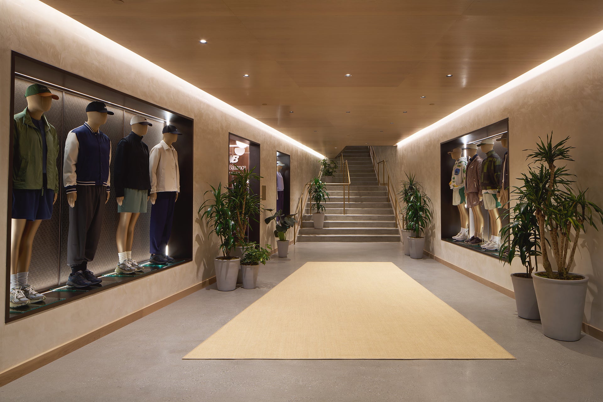 KITH store interior hallway with warm wood ceiling, lit display cases of mannequins in jackets and caps, potted palms and a central staircase