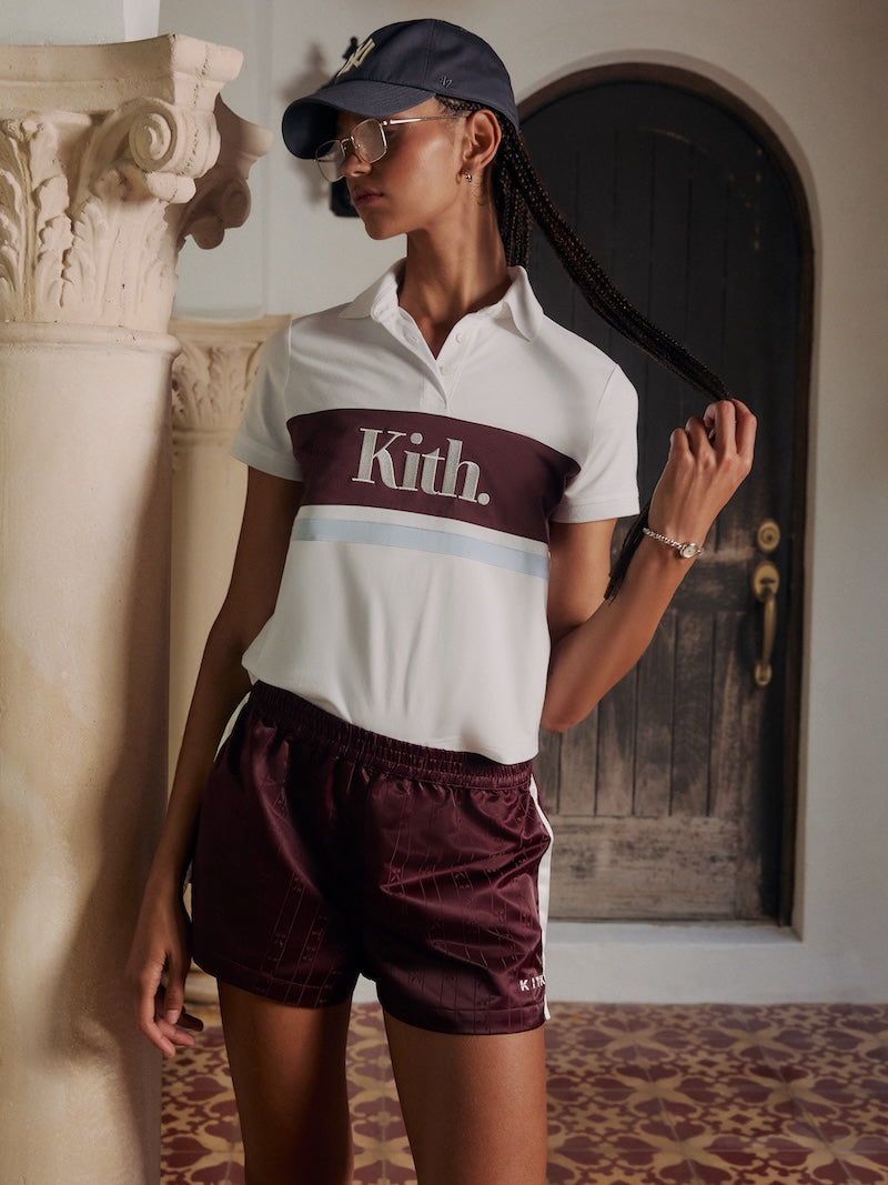 Woman by a stucco column in a navy Yankees cap, white and burgundy Kith colorblock polo shirt and burgundy monogram track shorts