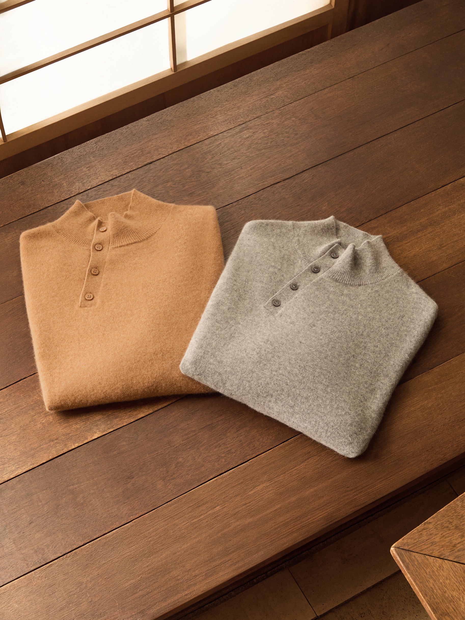 Two folded cashmere button mock-neck sweaters, camel and heather grey, flat-lay on dark wood floor